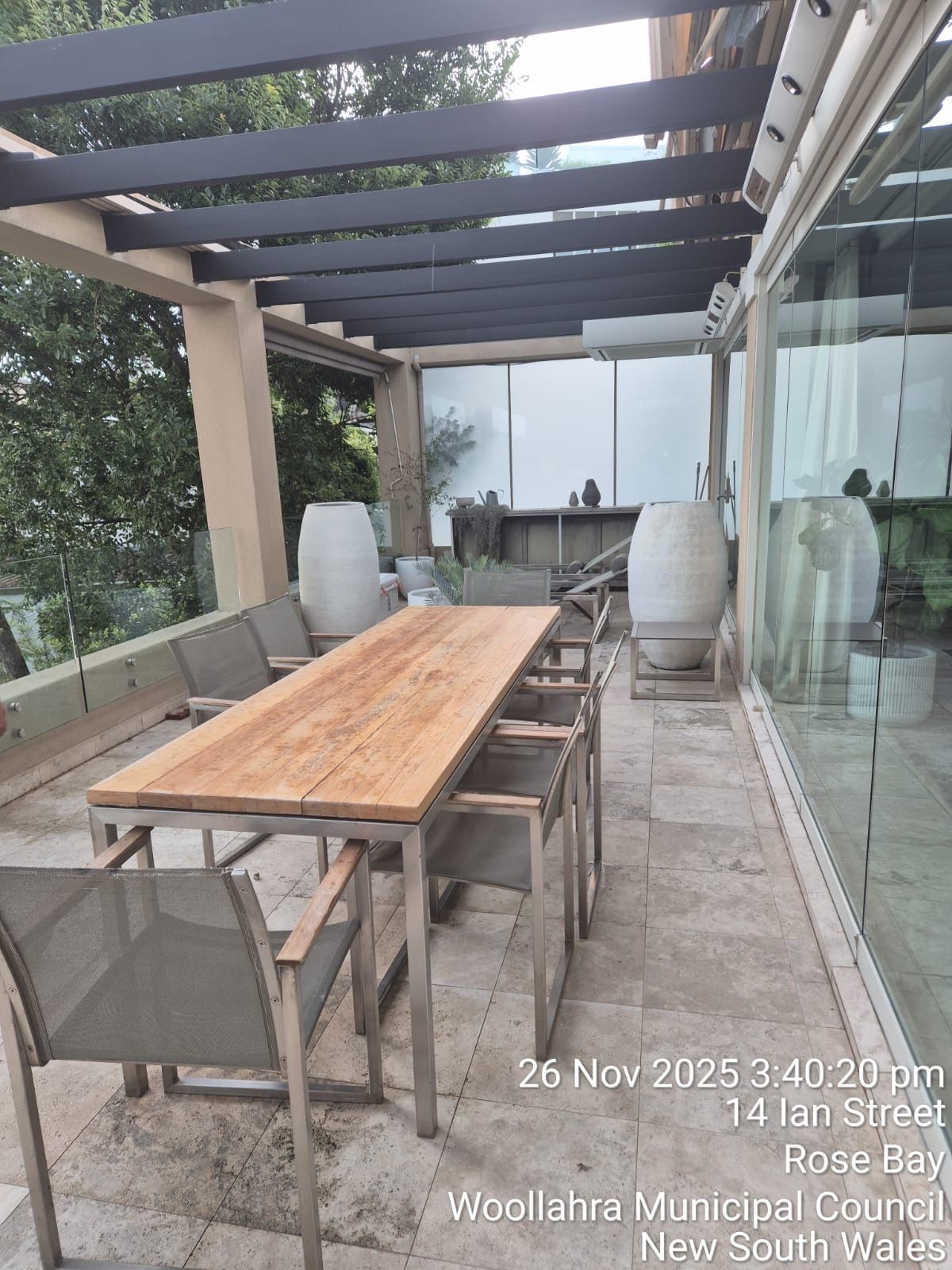 Outdoor patio with long wooden table, four chairs, glass wall, and pergola roof
