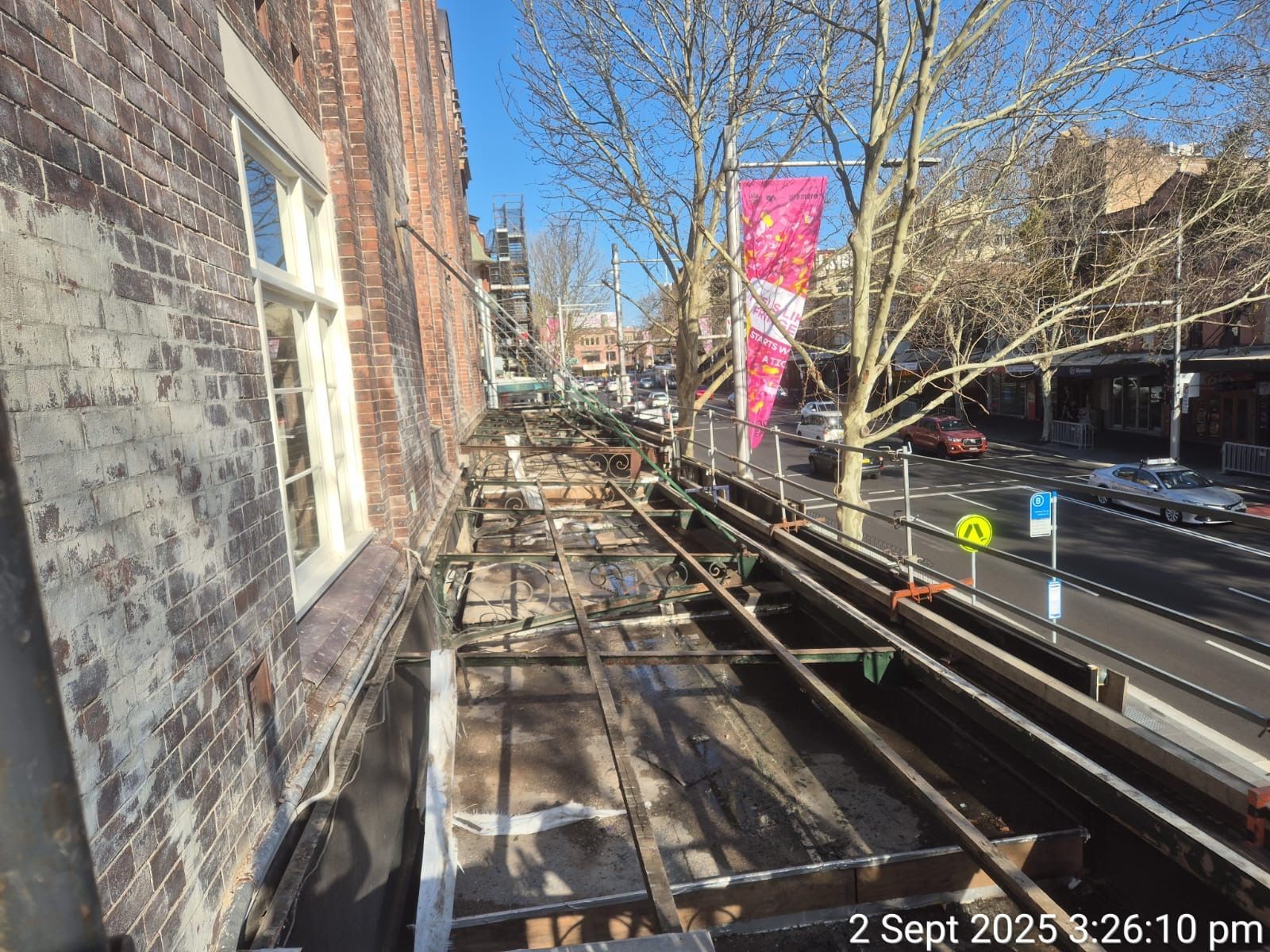Scaffolding along a brick building beside a road, with leafless trees and a pink banner on a sunny day.