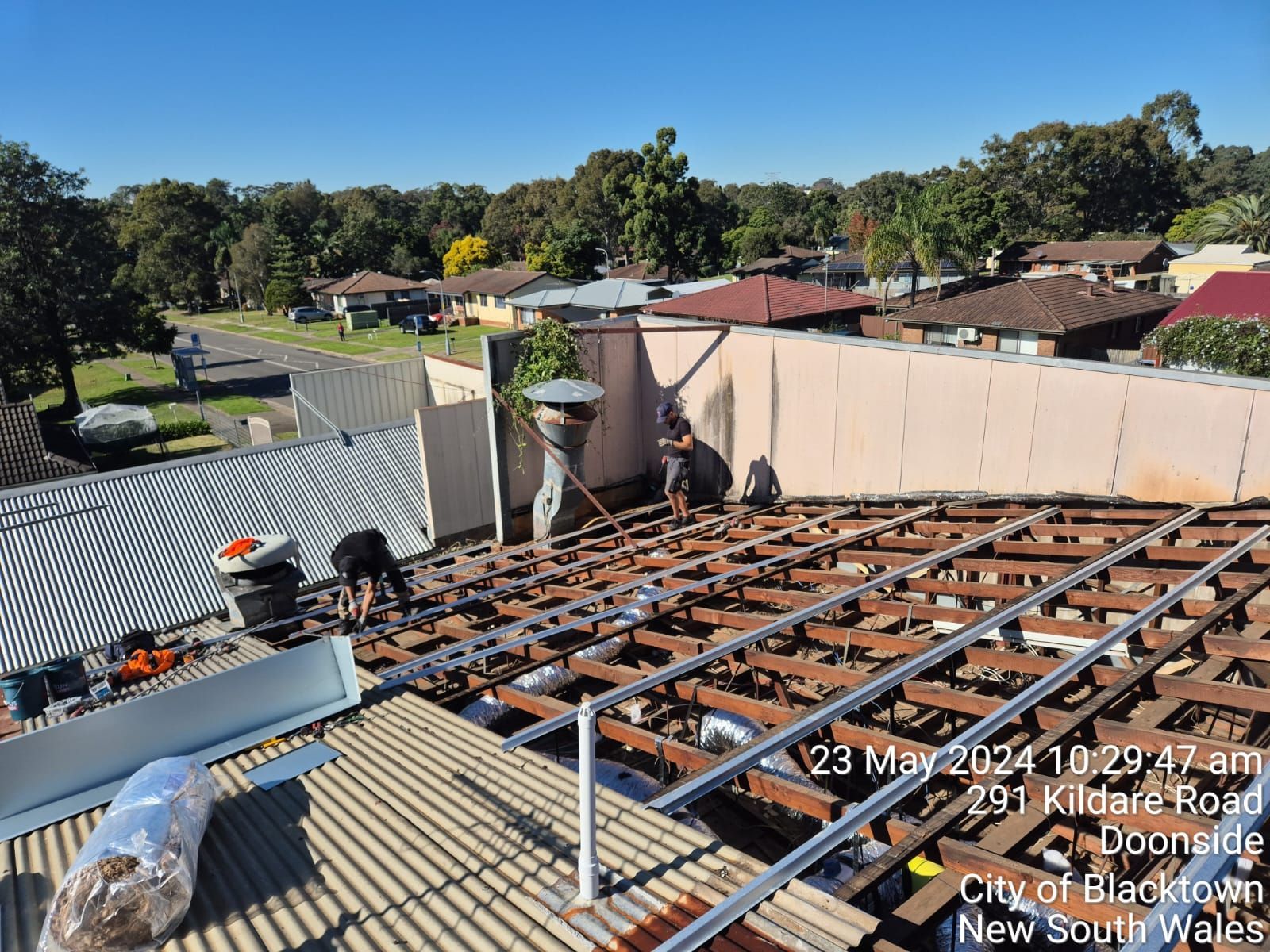 Rooftop under construction with exposed timber framing, tools, and suburban houses in the background.