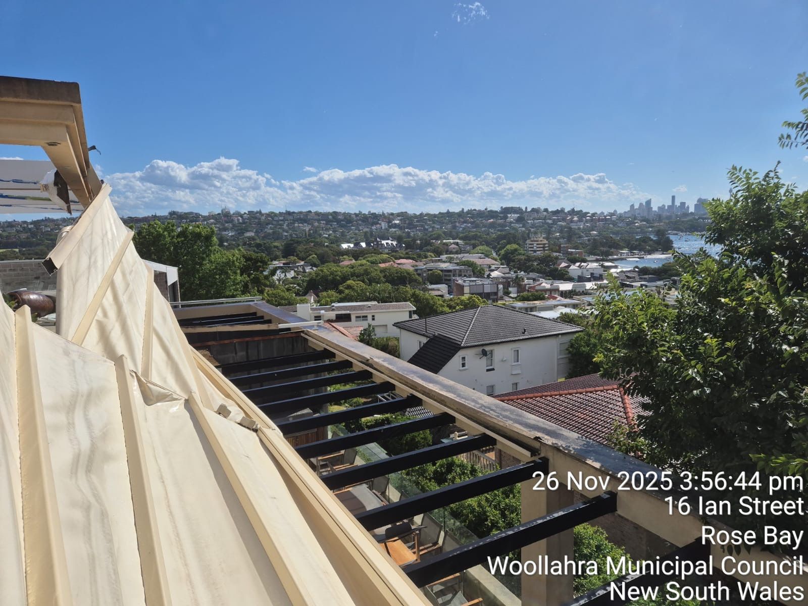 Rooftop view over a suburb with exposed beams and a white tarp in the foreground, under a cloudy sky