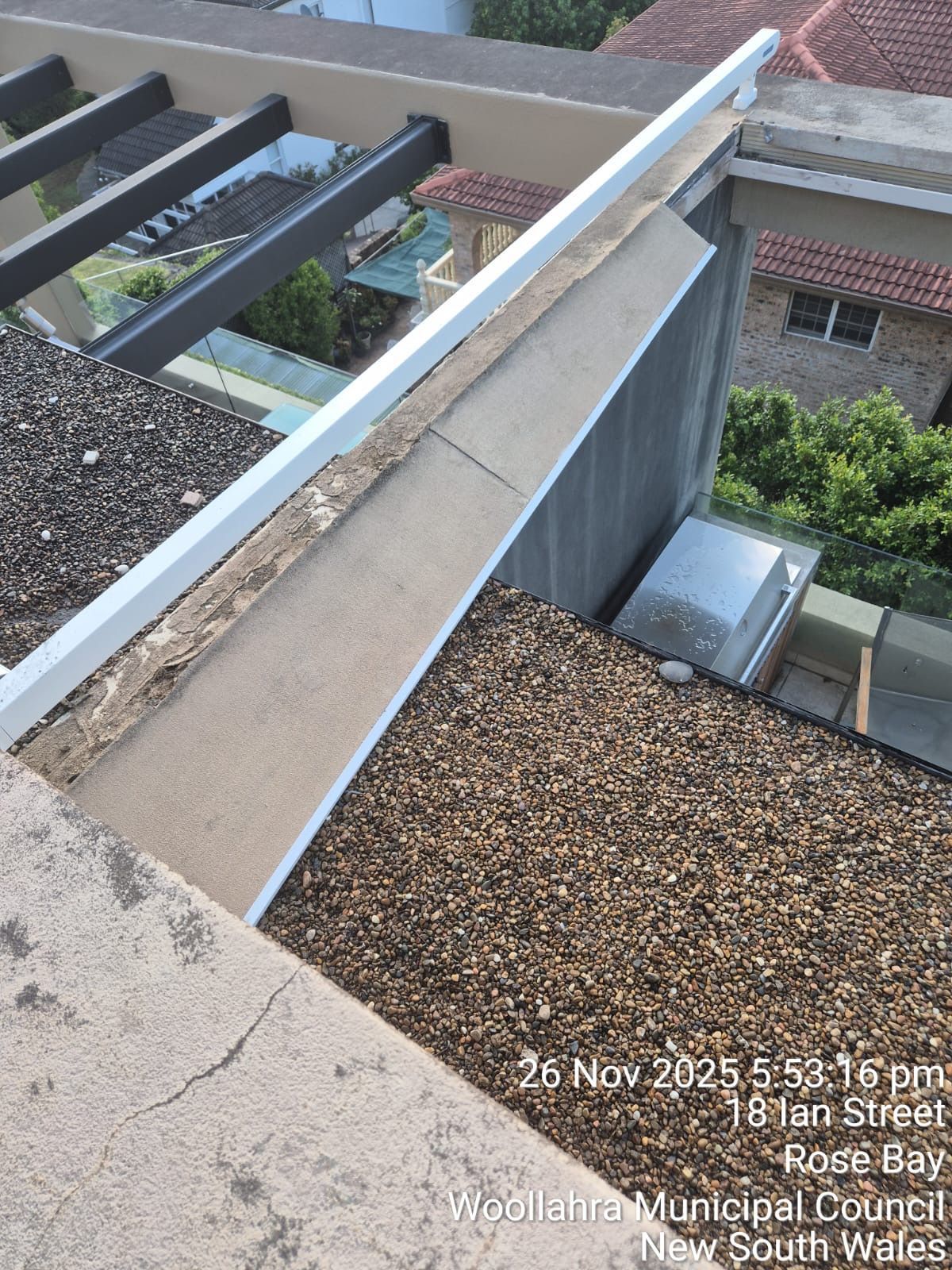 Rooftop with gravel, beams, and a concrete parapet overlooking nearby buildings and greenery