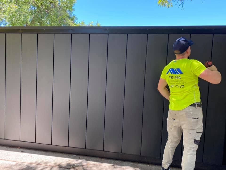 Worker in neon yellow shirt painting a dark fence outdoors under a clear sky
