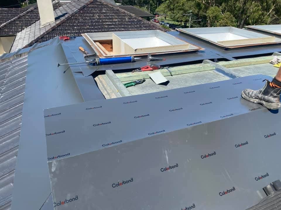 Roof repair with gray underlayment, blue trim, skylights, and a worker’s boot visible on the right
