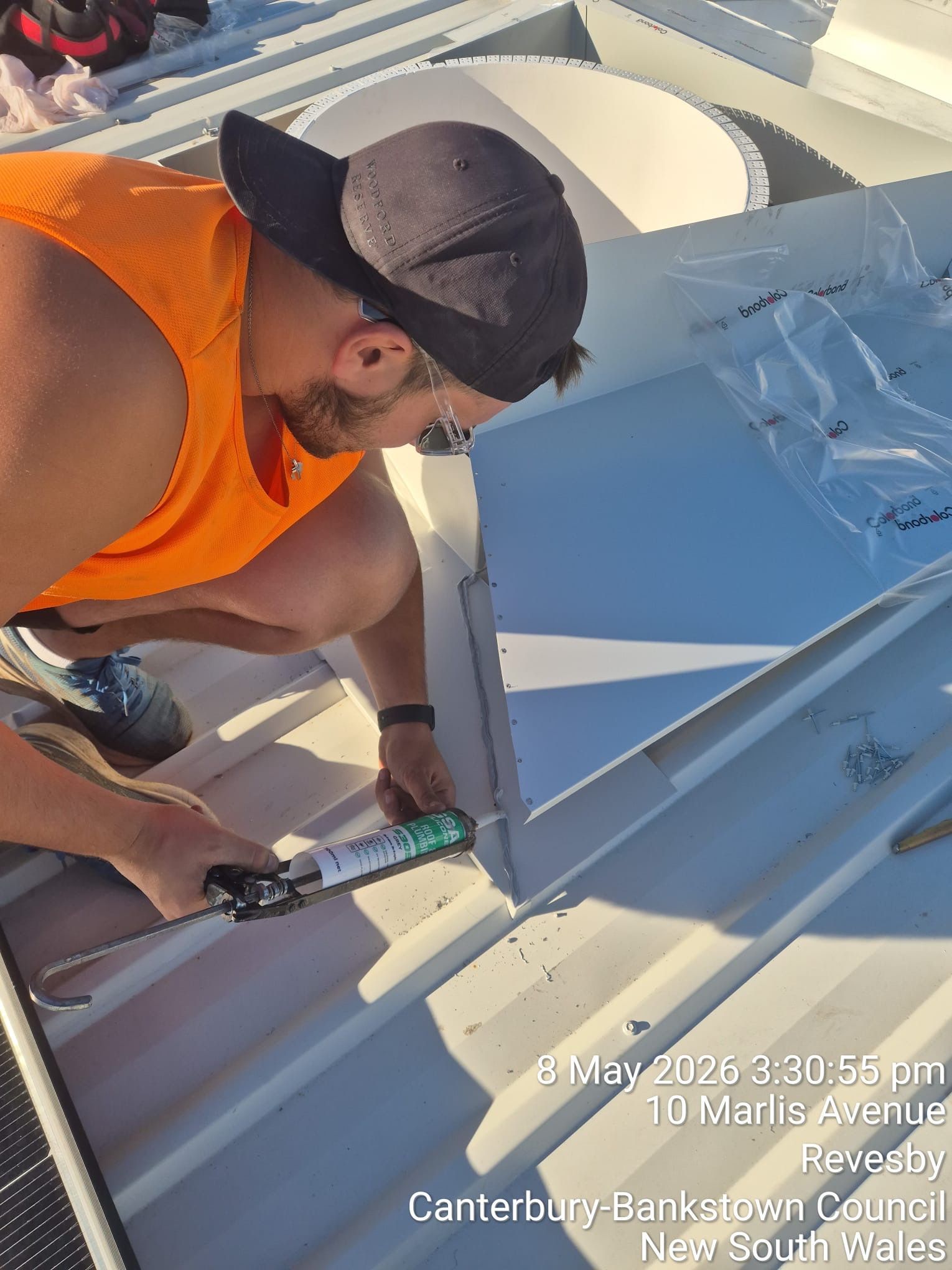 Worker in orange vest and black cap sealing a white rooftop seam with a caulking gun.