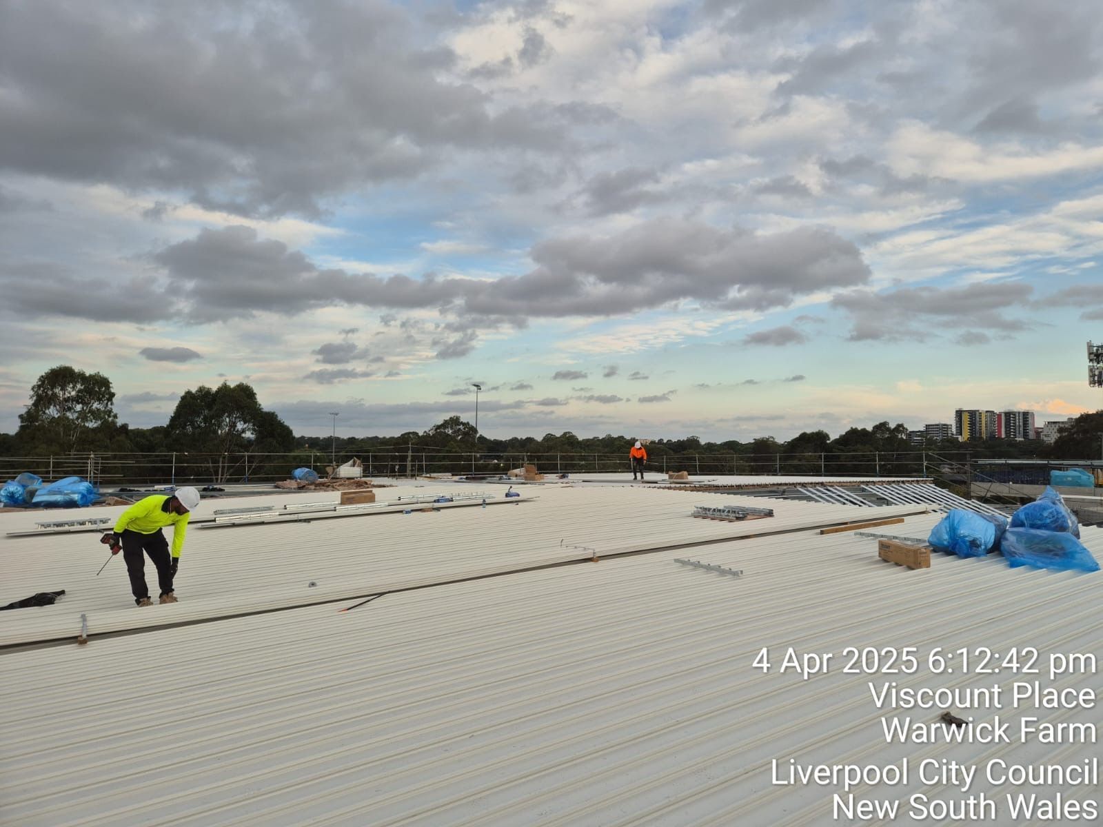 Workers on a large rooftop, spreading white roofing material under a cloudy sky.