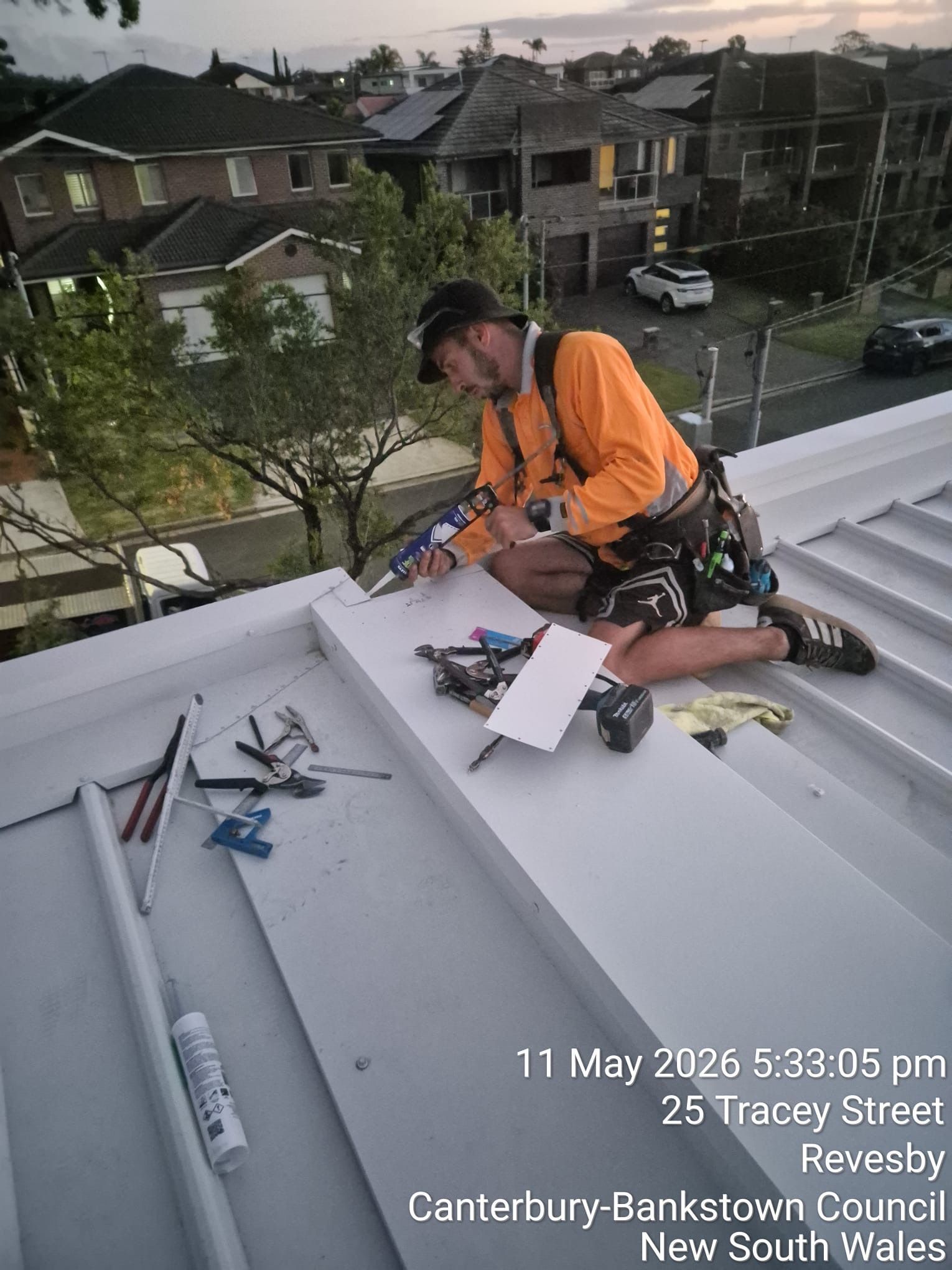 Worker in orange safety gear installing roofing on a steep white roof in a suburban neighborhood.