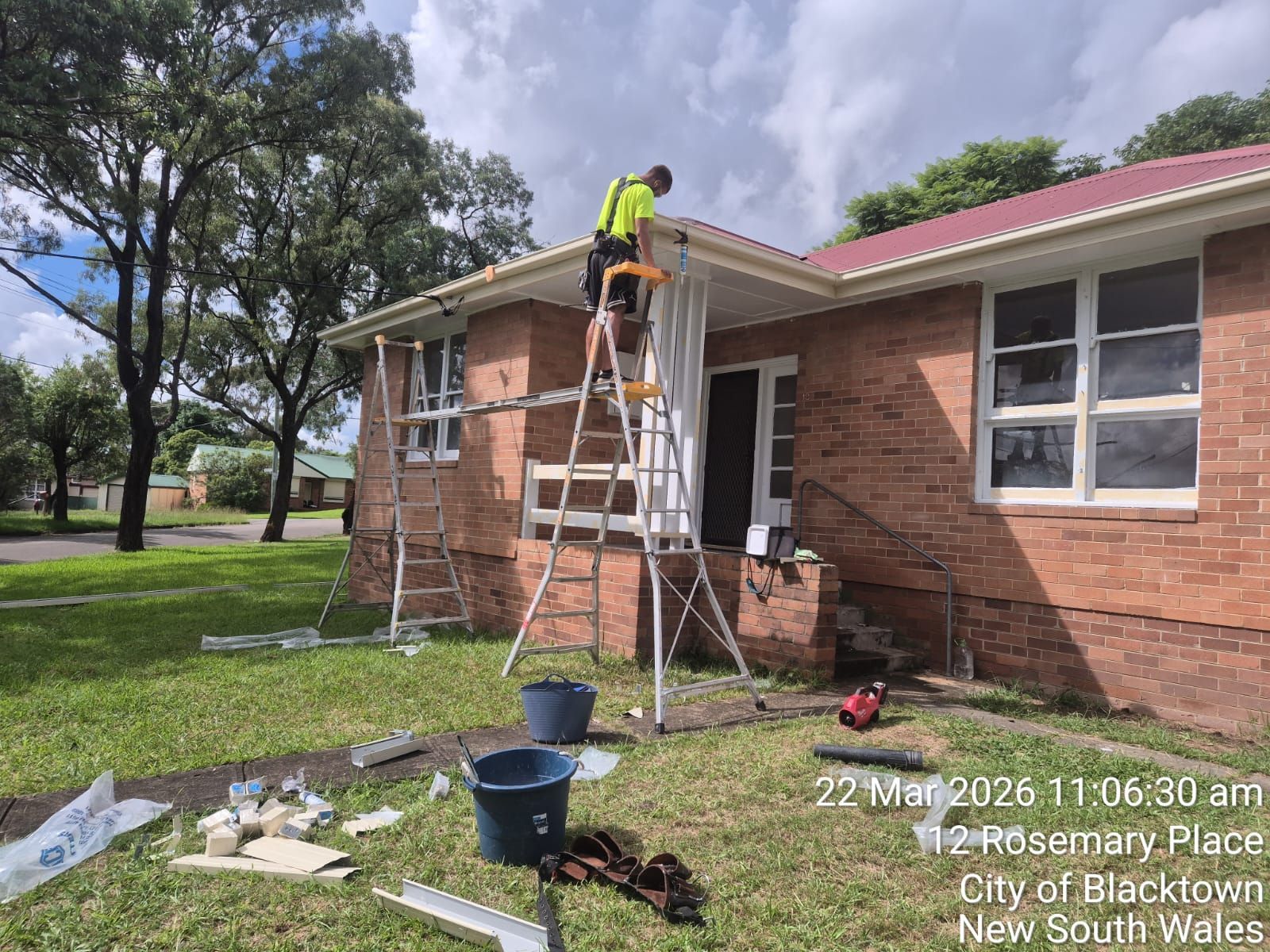 Worker on ladder repairing a brick house roof edge, with tools and debris on the lawn.