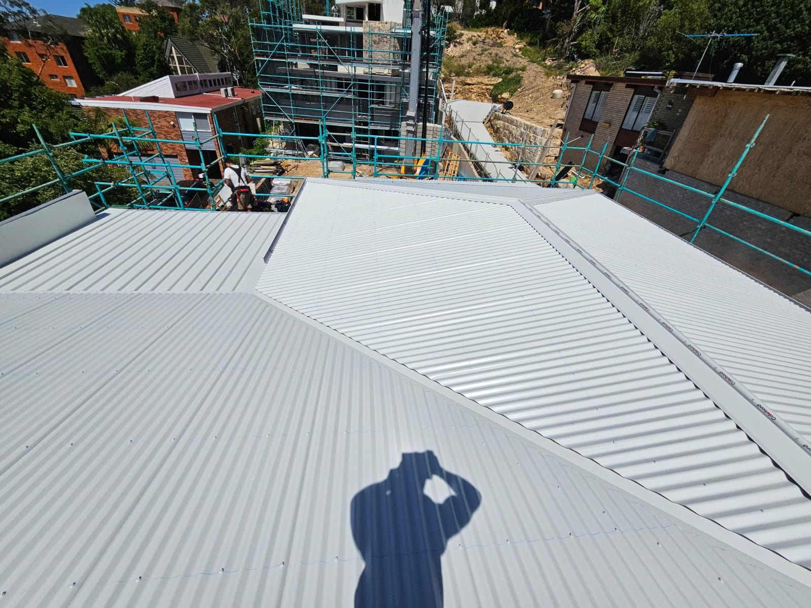 Rooftop with gray corrugated metal panels and a worker’s shadow, overlooking a building site below