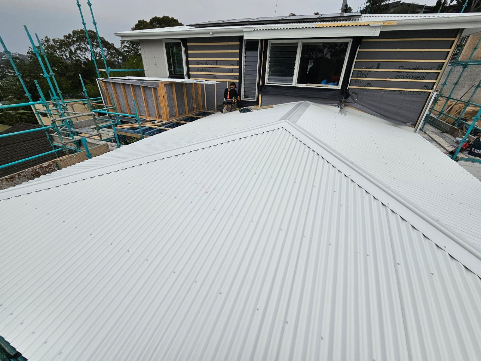 White corrugated metal roof under construction beside a house with scaffolding and open windows