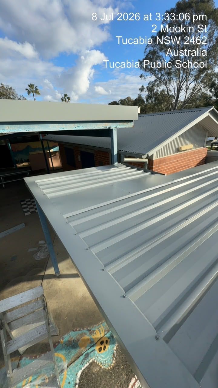 Corrugated metal roof beside a school building under a blue sky, with a sign in the background