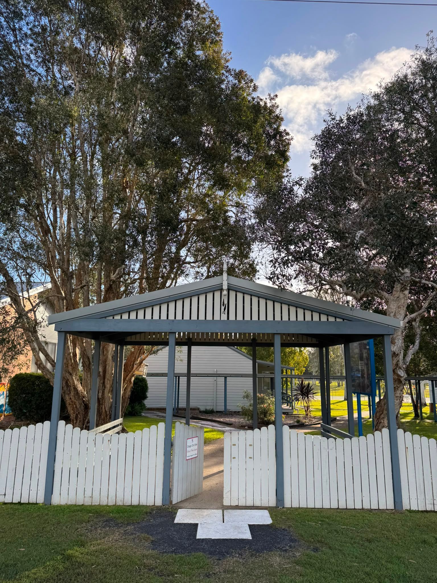 White gazebo with a picket fence in a grassy yard, framed by tall trees and a partly cloudy sky