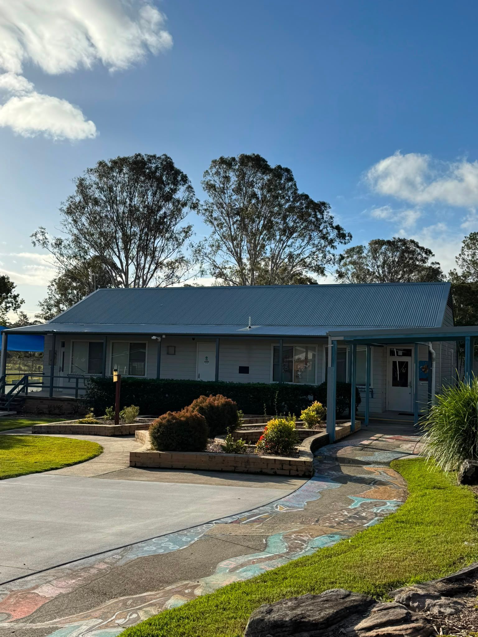 Single-story house with metal roof, front porch, garden bed, and curved driveway under a blue sky