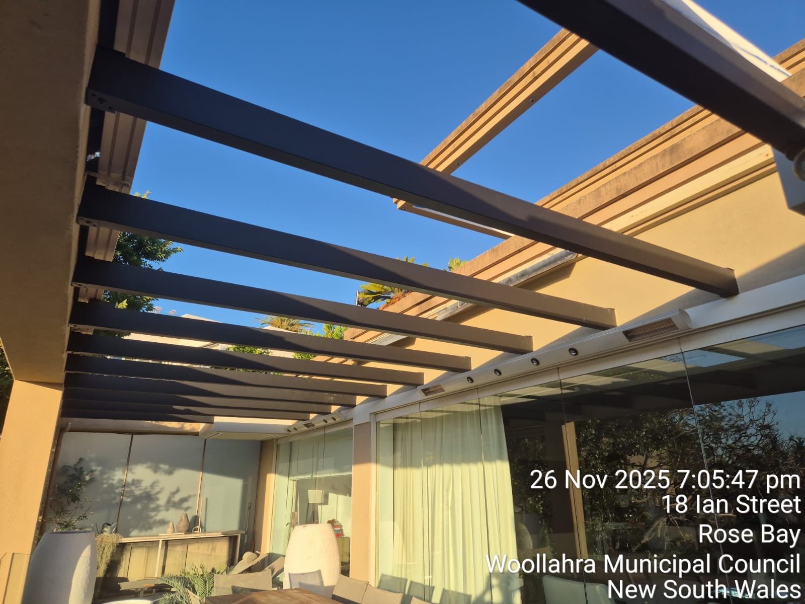 Patio with dark wooden pergola beams, beige walls, and clear blue sky at sunset