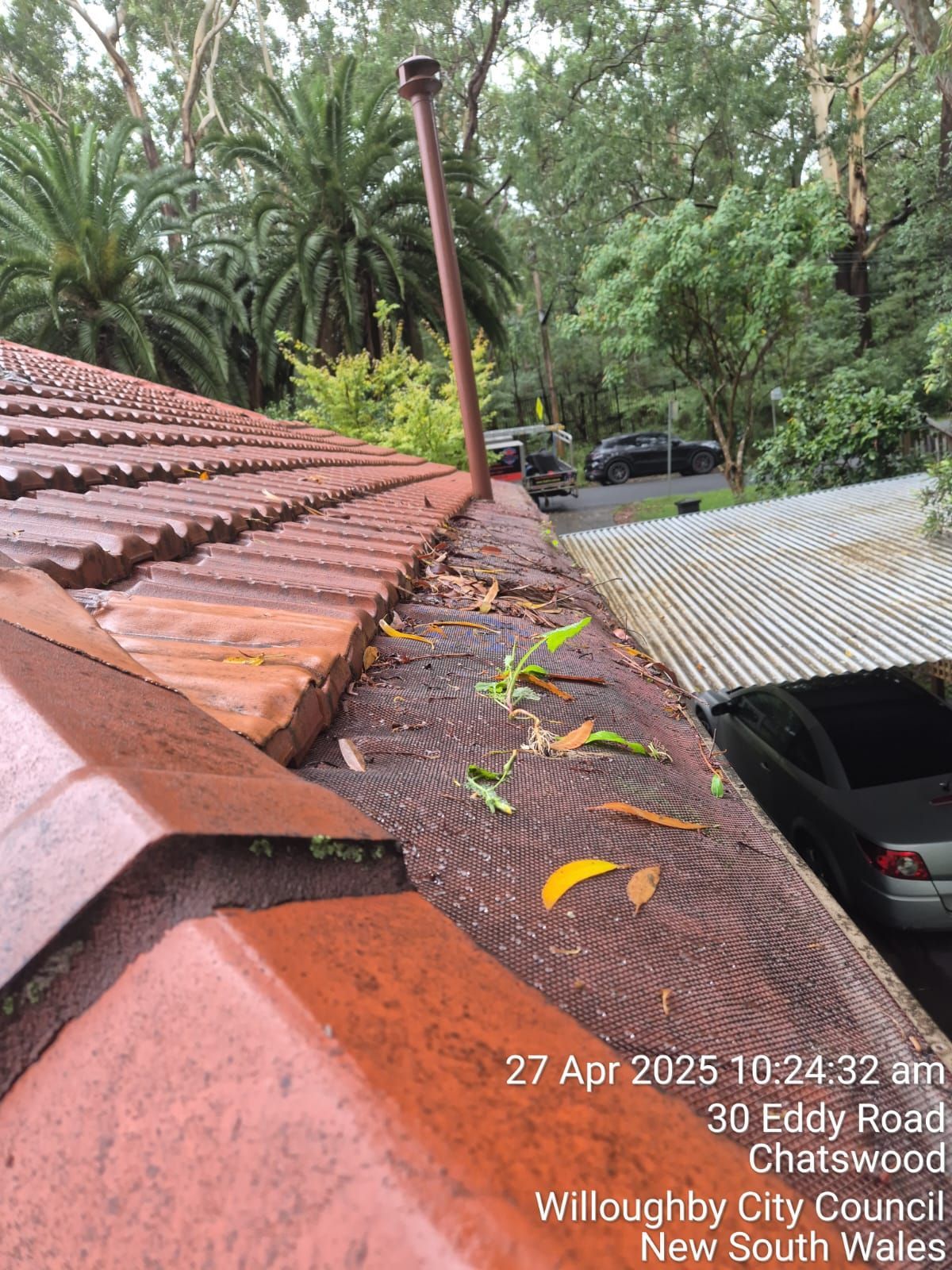 Red tiled roof with leaves and gutter, overlooking a corrugated roof and parked car by trees