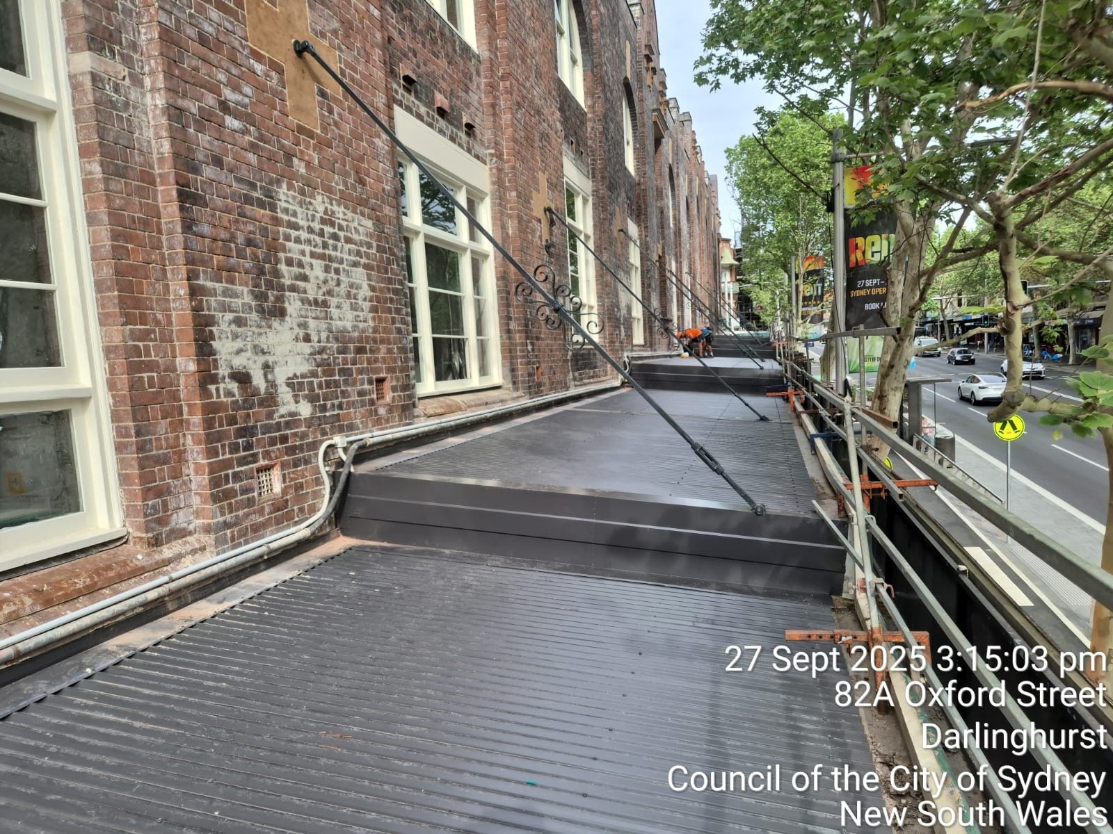 Scaffolding walkway beside a brick building on a tree-lined street with parked cars