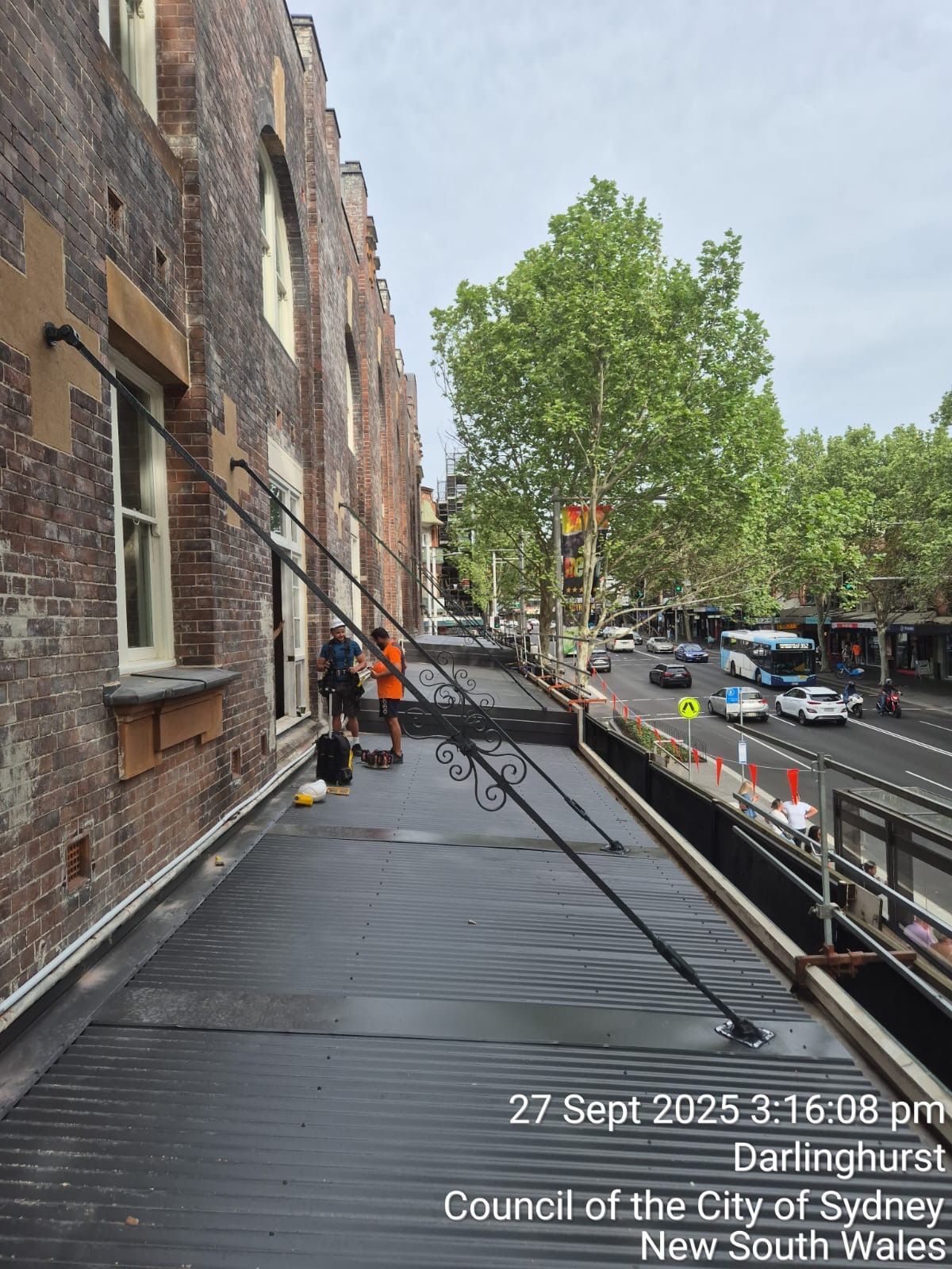 Worker on a rooftop walkway beside brick buildings, with trees and a street below.