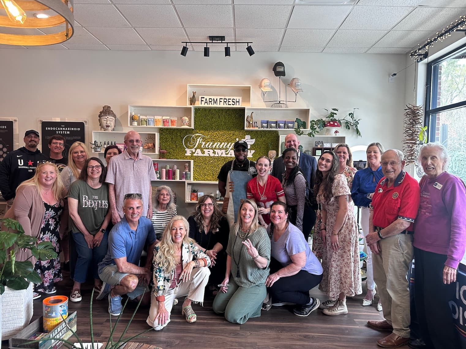 Large group posing indoors in front of a plant display and shelf, smiling at the camera.
