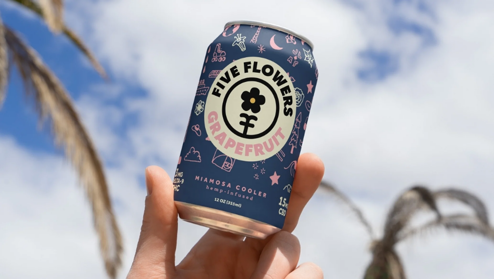 Hand holding a can of Five Flowers grapefruit hemp infused sparkling water against a blue sky with palm trees.