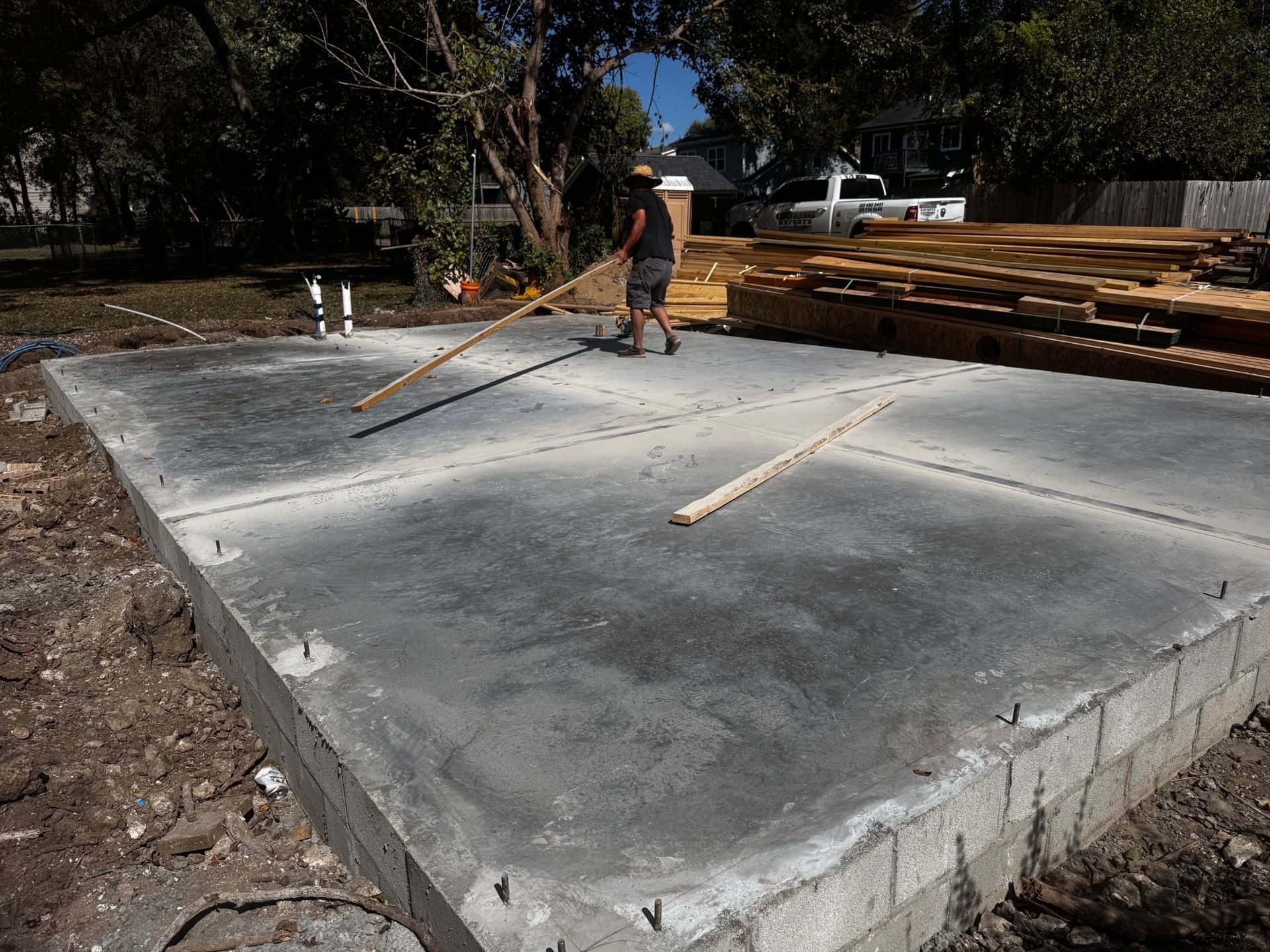 Worker smoothing a concrete slab beside stacked lumber in an outdoor construction site.