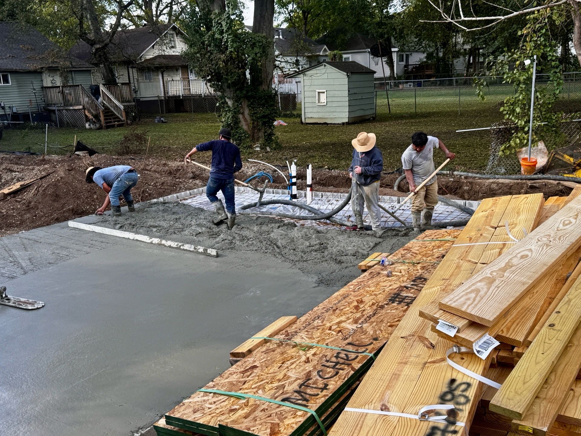 Workers pouring a concrete slab beside stacked lumber at a residential construction site