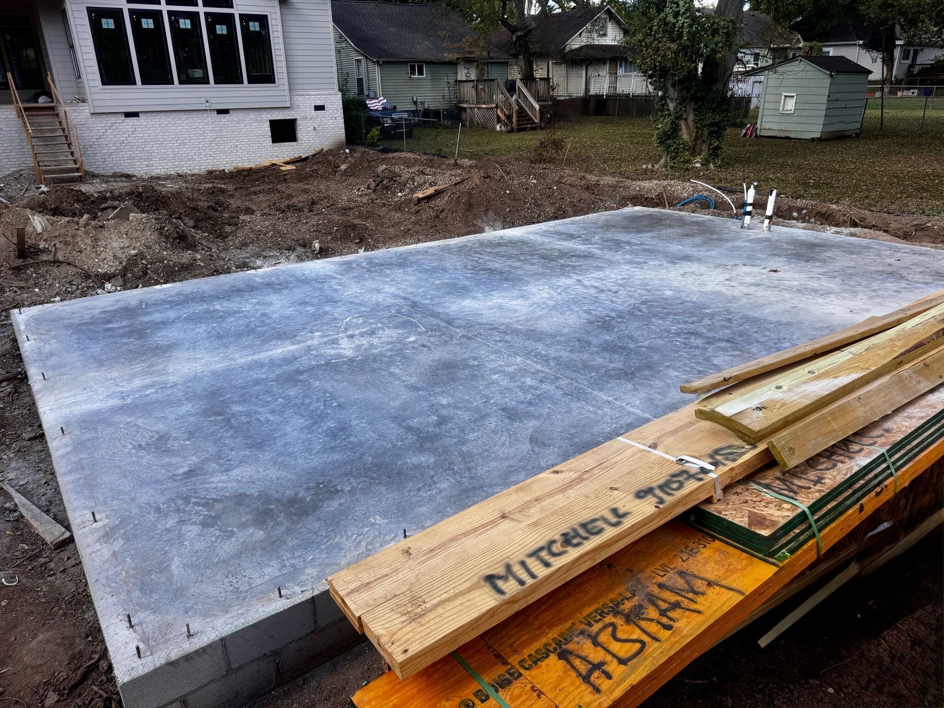 Concrete slab foundation under construction with lumber stacked in the foreground and a house nearby