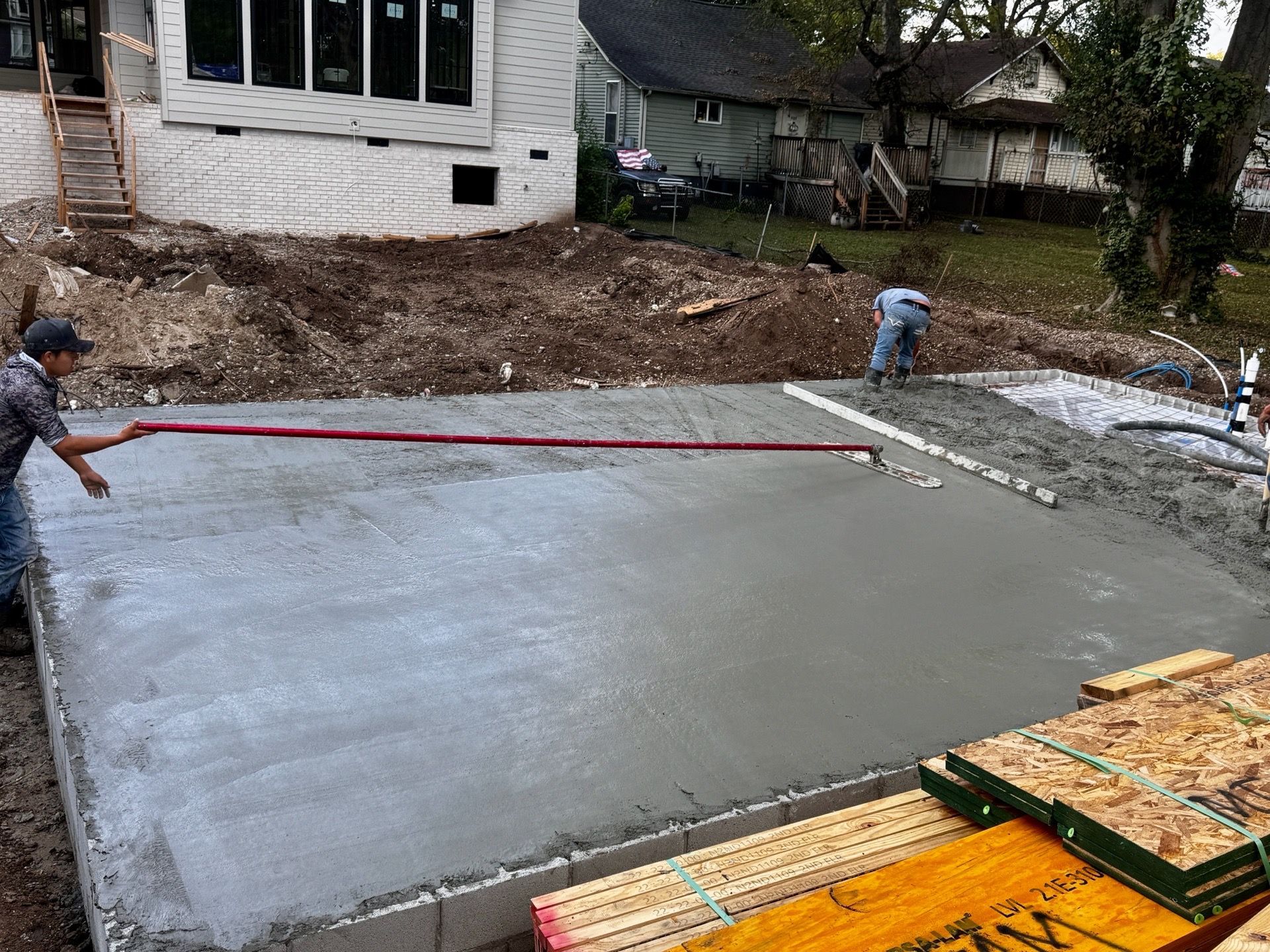 Workers smoothing a fresh concrete slab beside a house, with a long screed and construction materials nearby