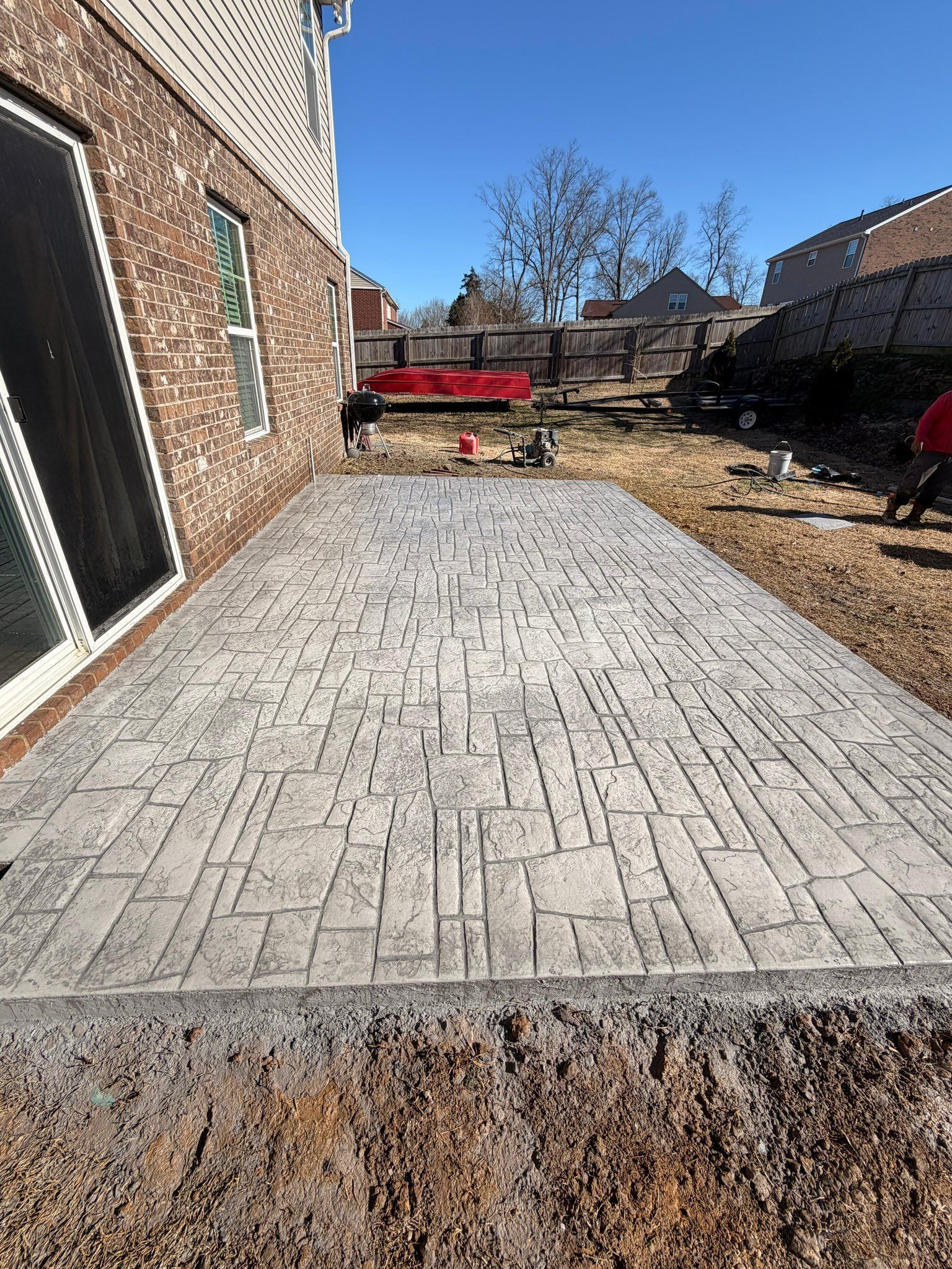 Backyard with new light-gray brick patio beside a house, red truck in the distance, and bare trees under blue sky