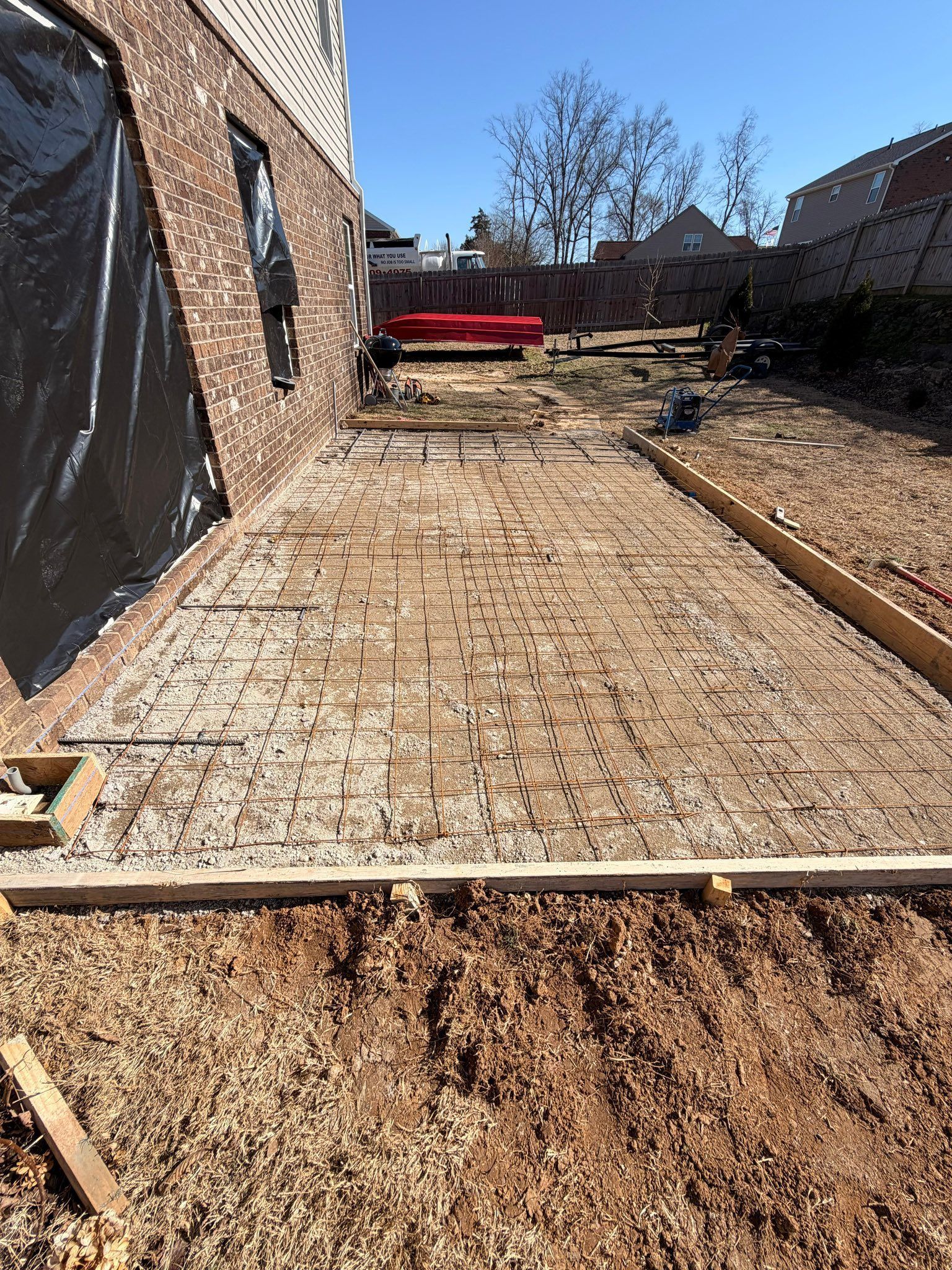 Backyard construction site with a gravel path, wood forms, brick house wall, and red wheelbarrow.