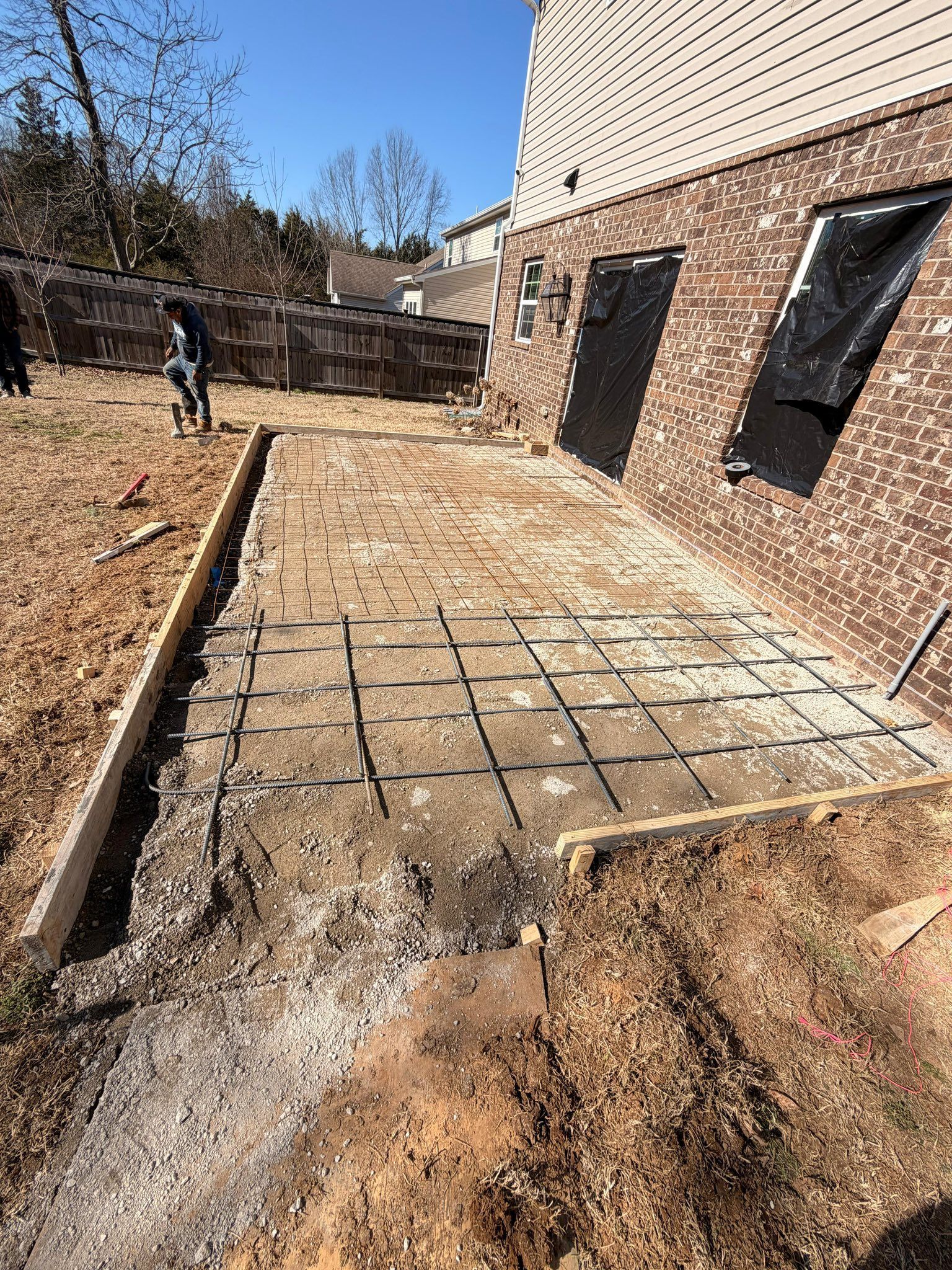 Backyard patio under construction beside a house, with rebar grid and muddy ground.