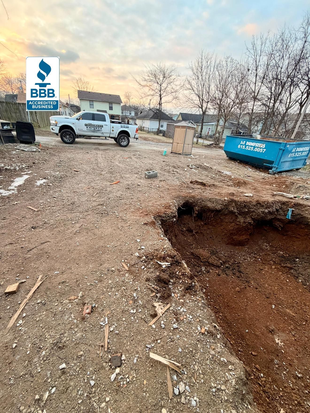 Dirt lot with a white truck, blue dumpster, BBB sign, and a large excavation hole in the foreground