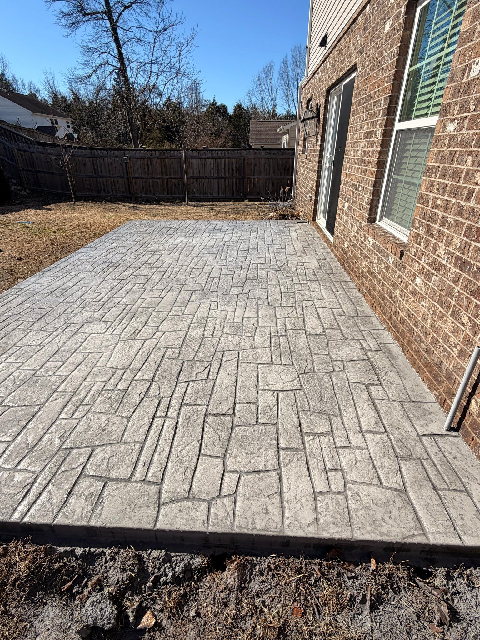 Brick patio in a backyard beside a house, with a wooden fence and leafless trees in the background.