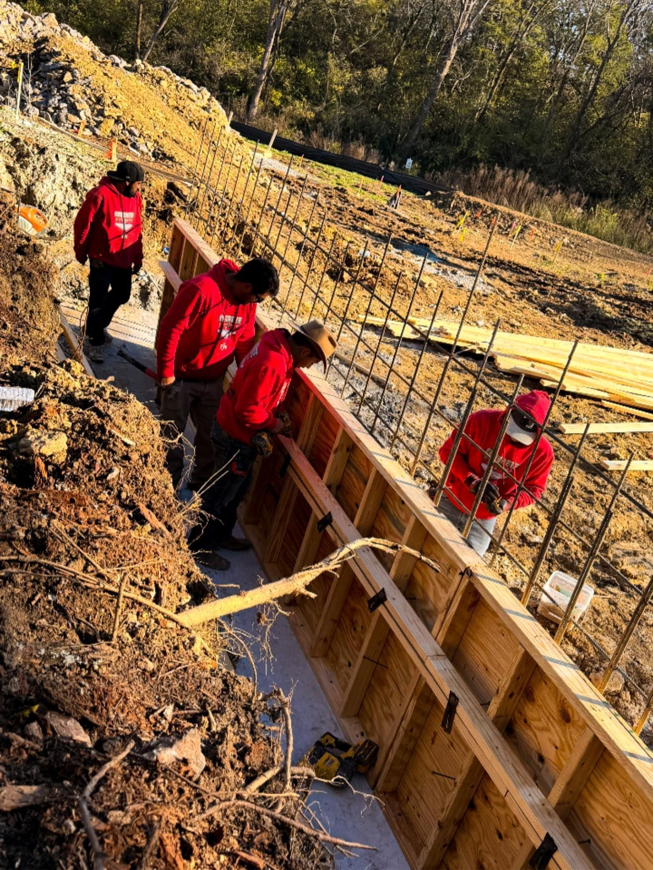 Workers in red jackets on a dirt construction site beside wooden forms and rebar.