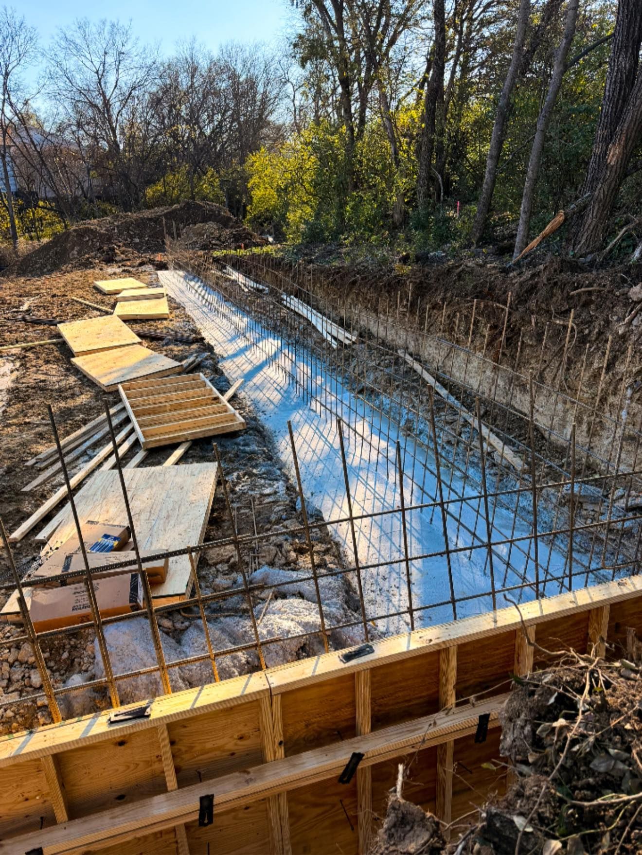 Concrete foundation under construction beside a muddy trench and wooded area, with rebar and wooden forms visible