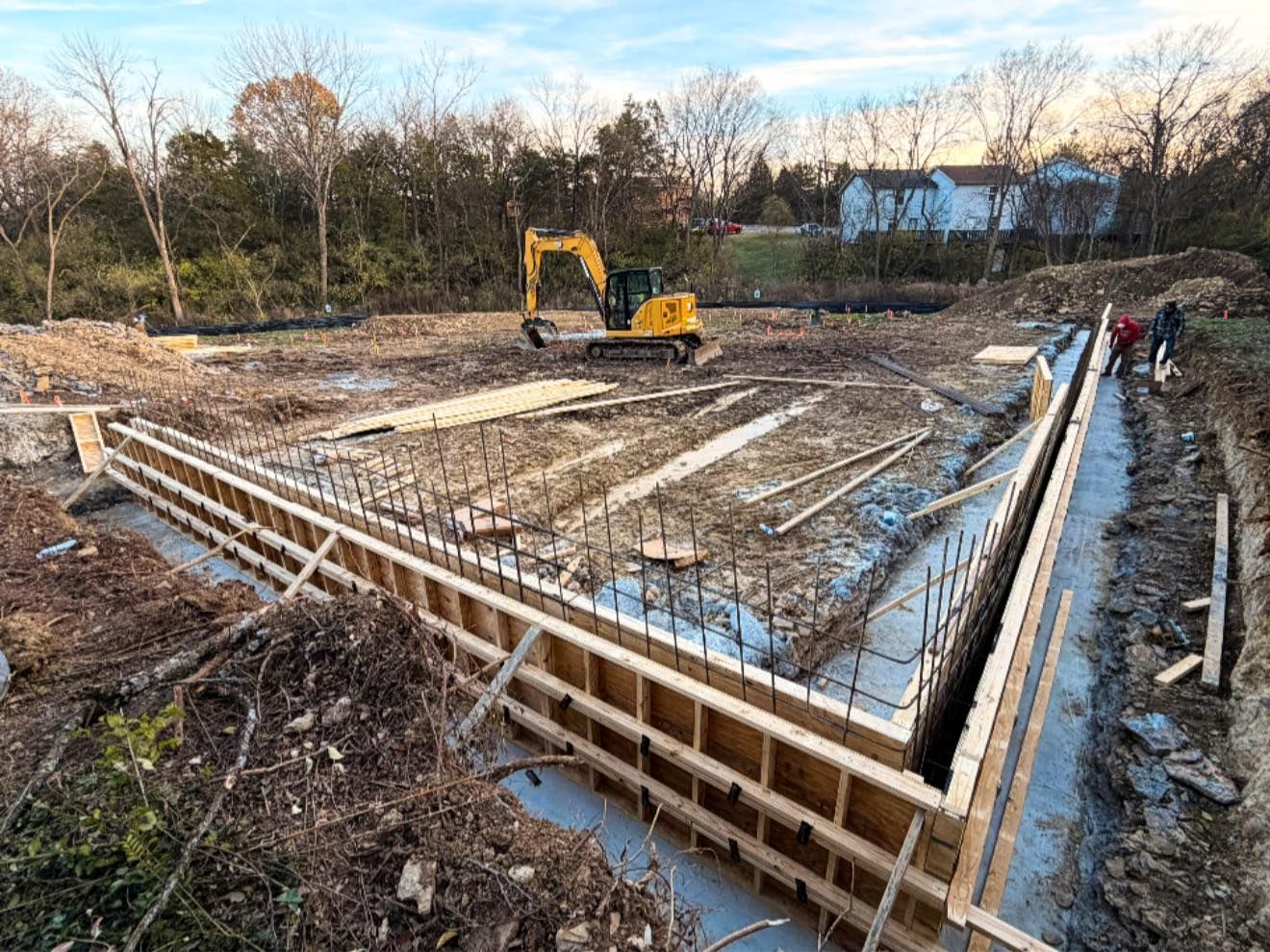 Concrete foundation construction site with excavator, wooden forms, and workers beside muddy trenches