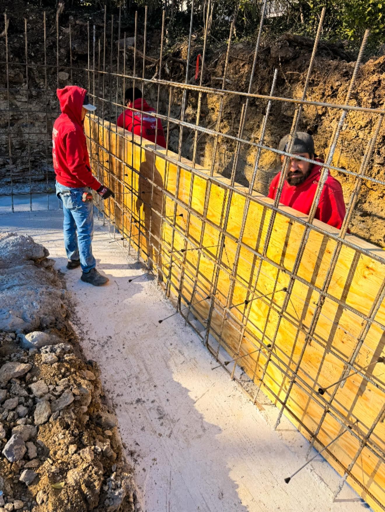 Two workers in red hoodies align a yellow retaining wall panel along a wooded dirt trench.