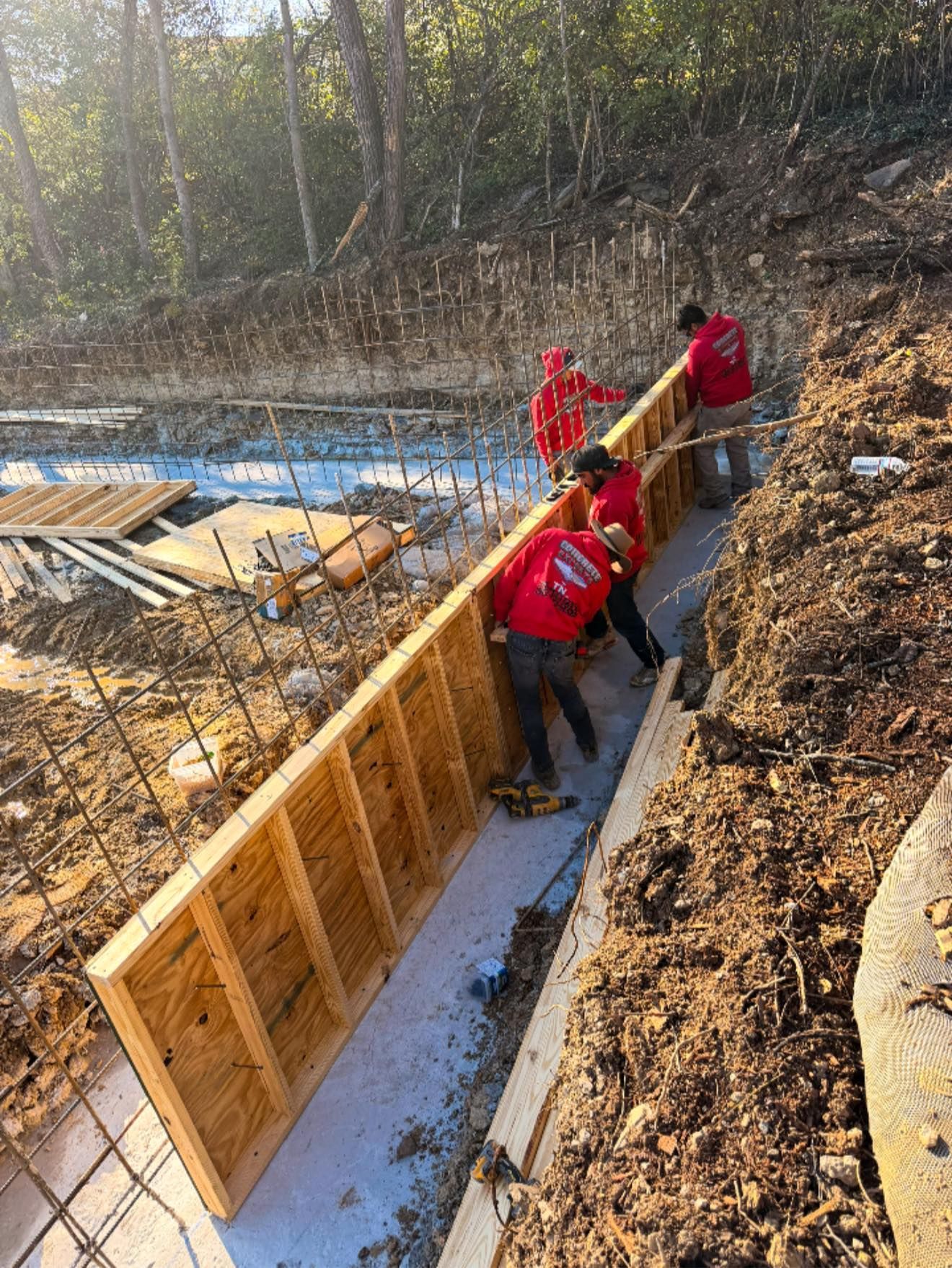 Workers in red jackets building a wooden retaining wall along a snowy wooded hillside