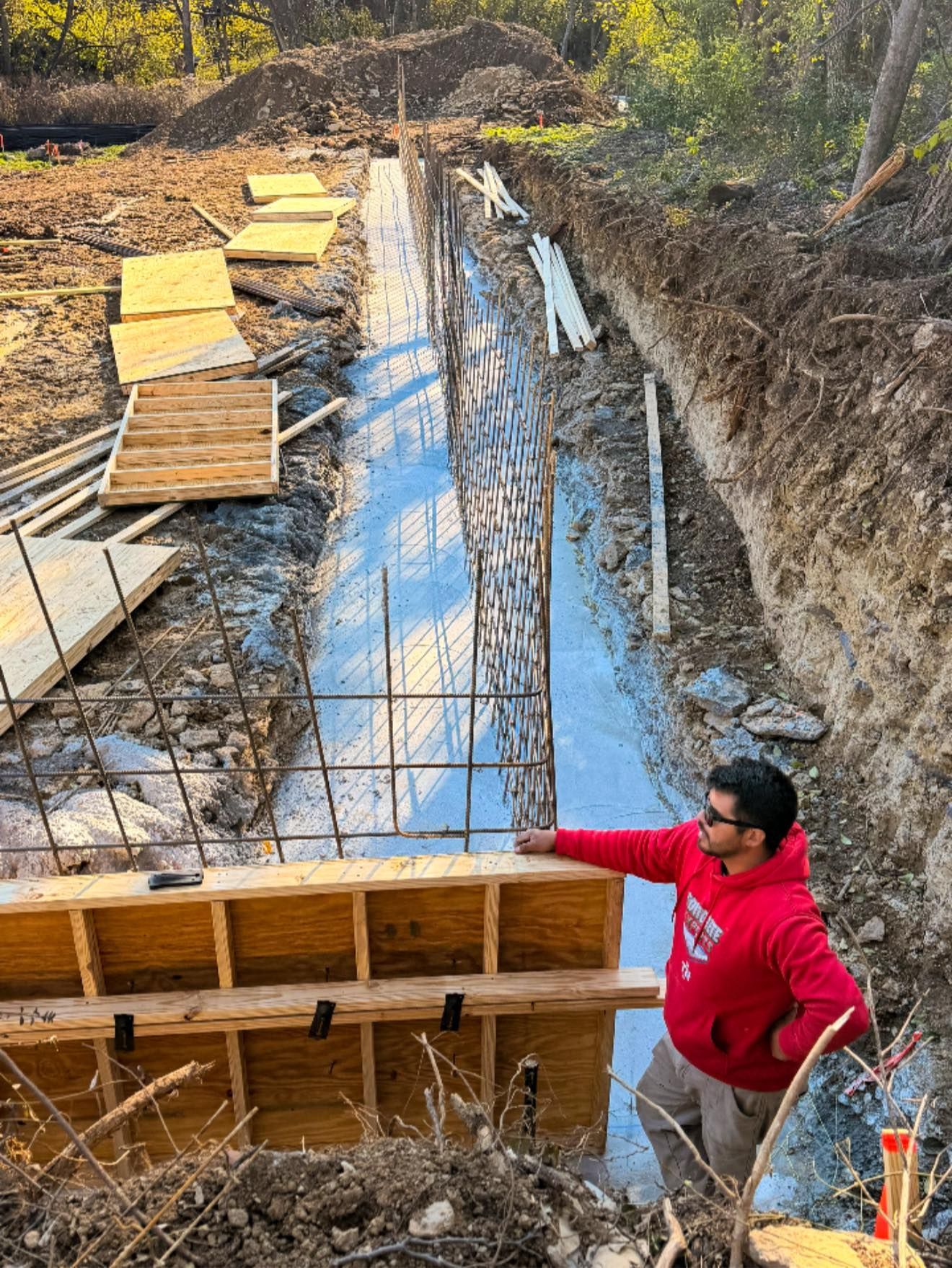 Worker in red hoodie points at wooden formwork around a long concrete trench on a construction site.
