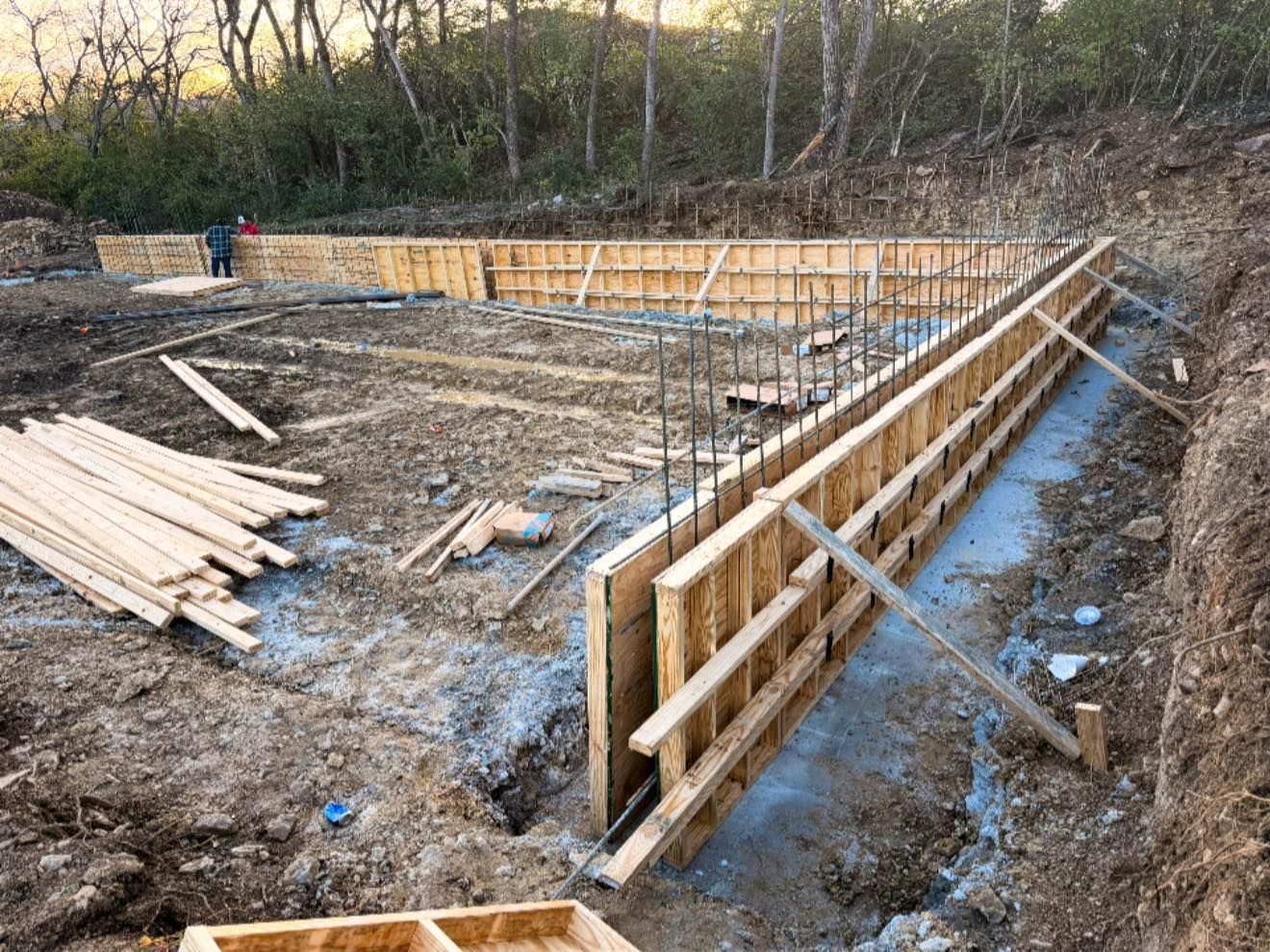 Wooden foundation forms and rebar at a construction site, with lumber stacked nearby and trees in the background