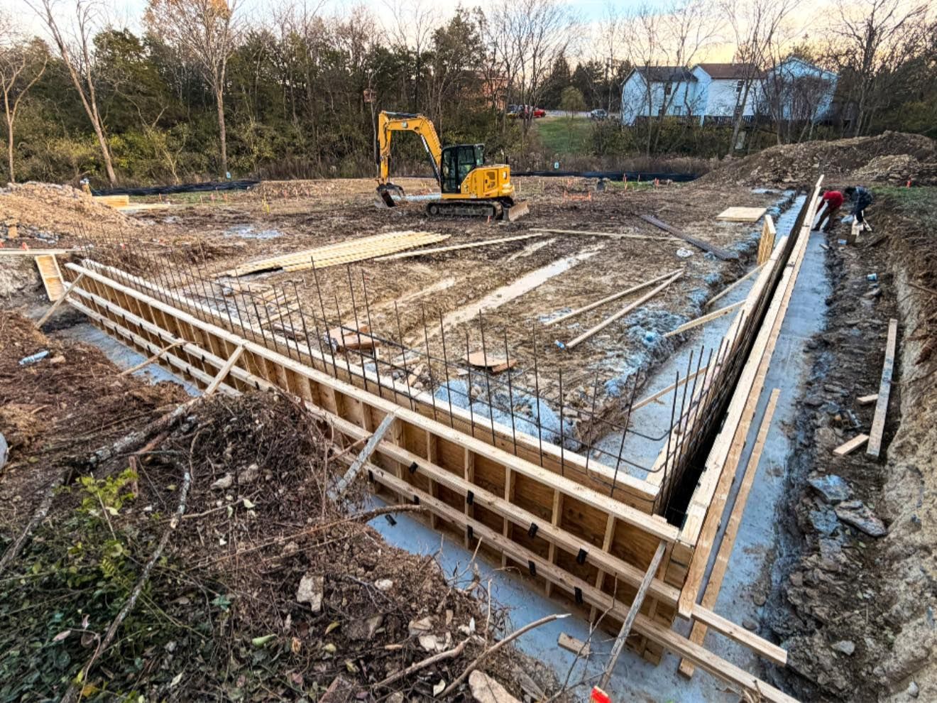 Construction site with excavator and concrete foundation forms in a muddy, wooded area