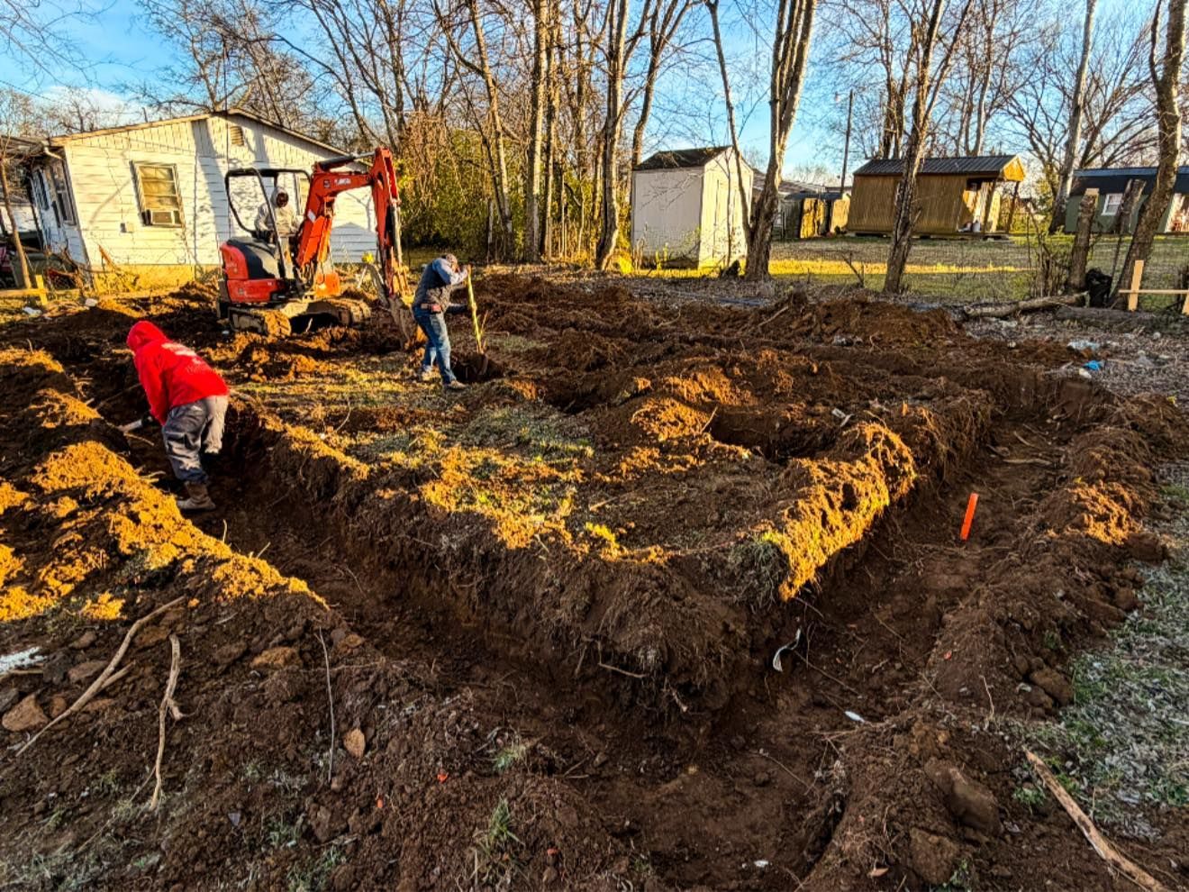 Workers digging a large muddy foundation trench beside a house and excavator on a sunny day