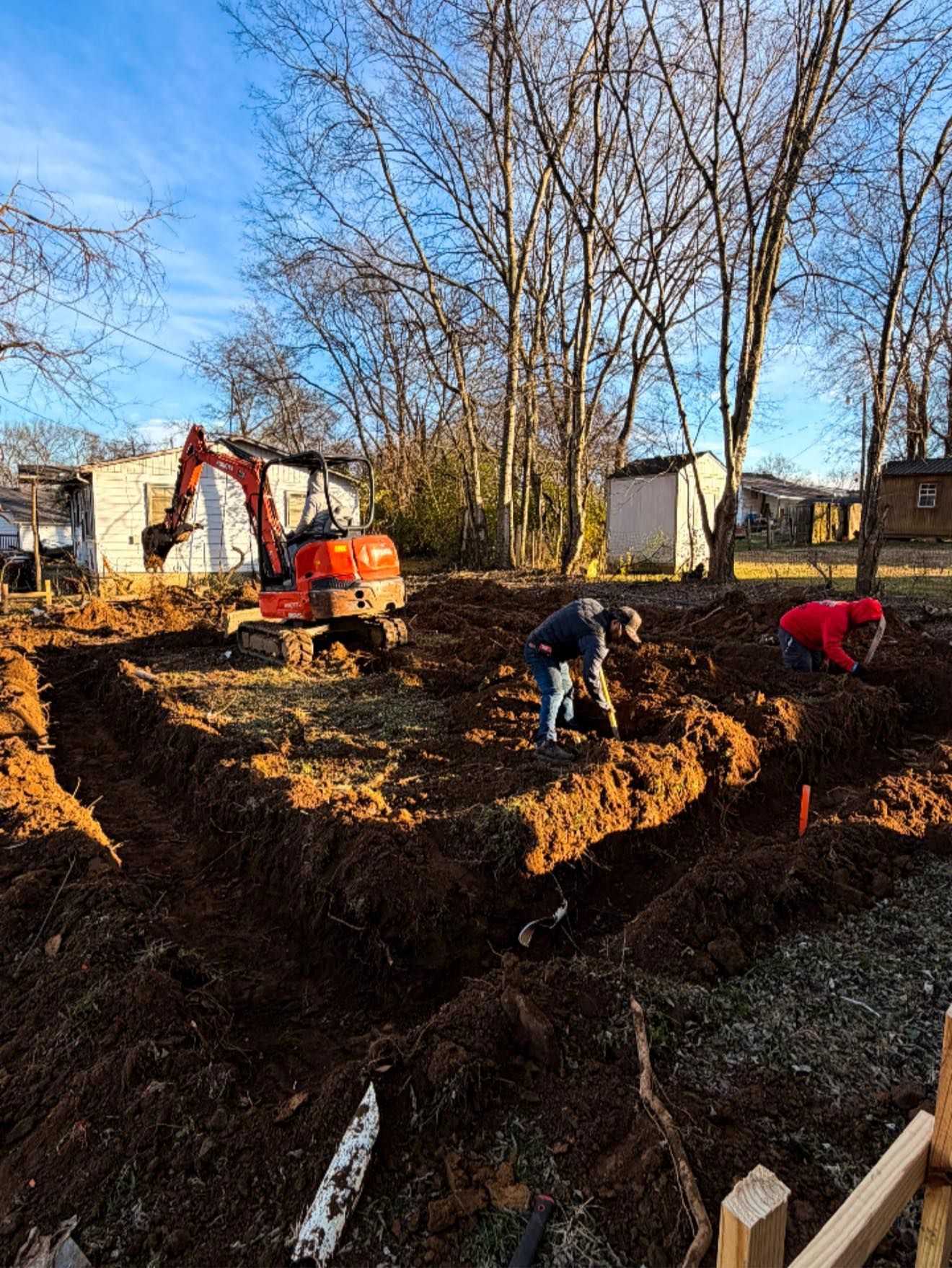 Workers digging a large trench at a construction site with an excavator and leafless trees in the background