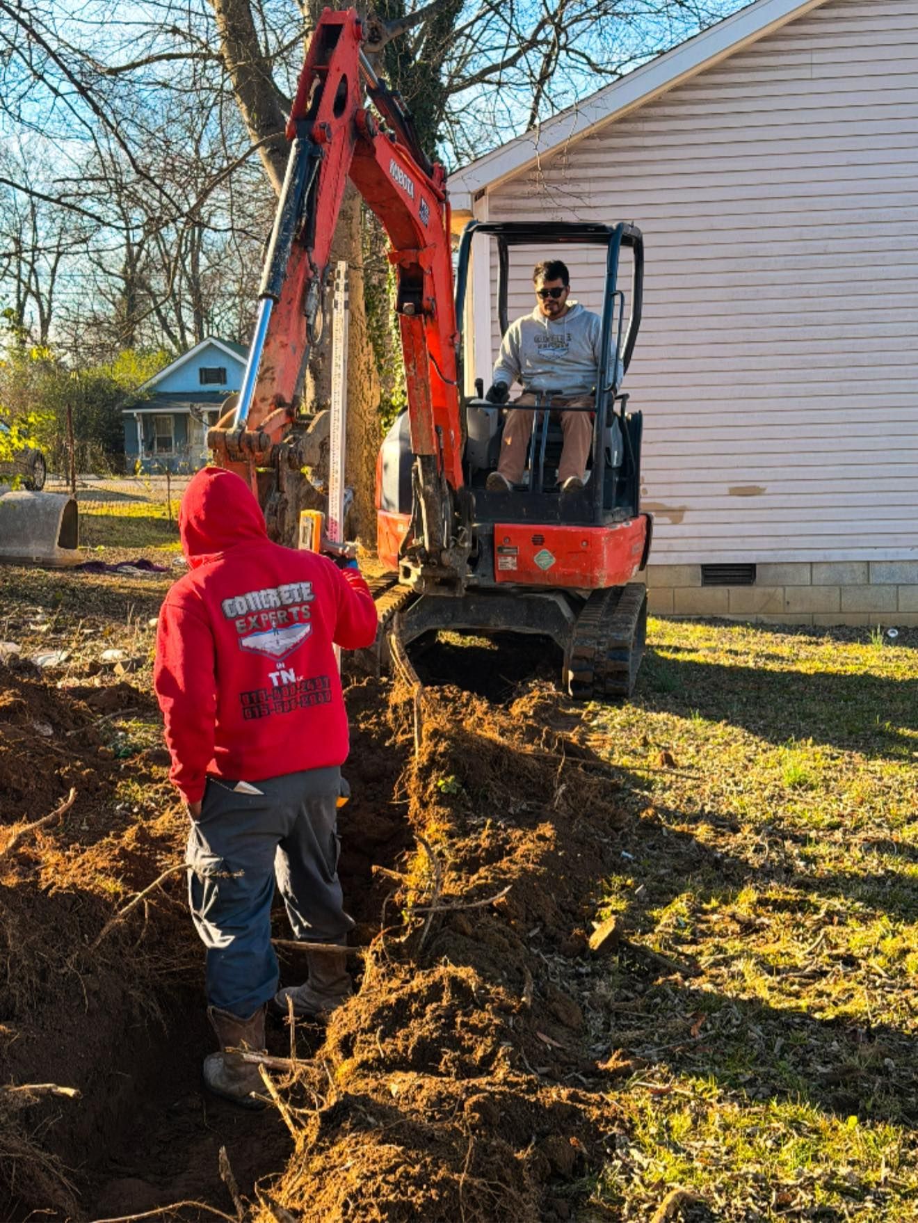 Worker operating a red excavator beside a house while another person stands nearby on a dirt trench.