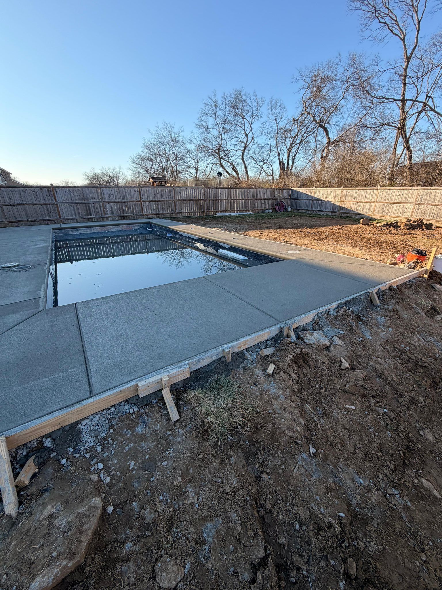 Outdoor concrete pool foundation with a small water-filled section and leafless trees under a clear blue sky