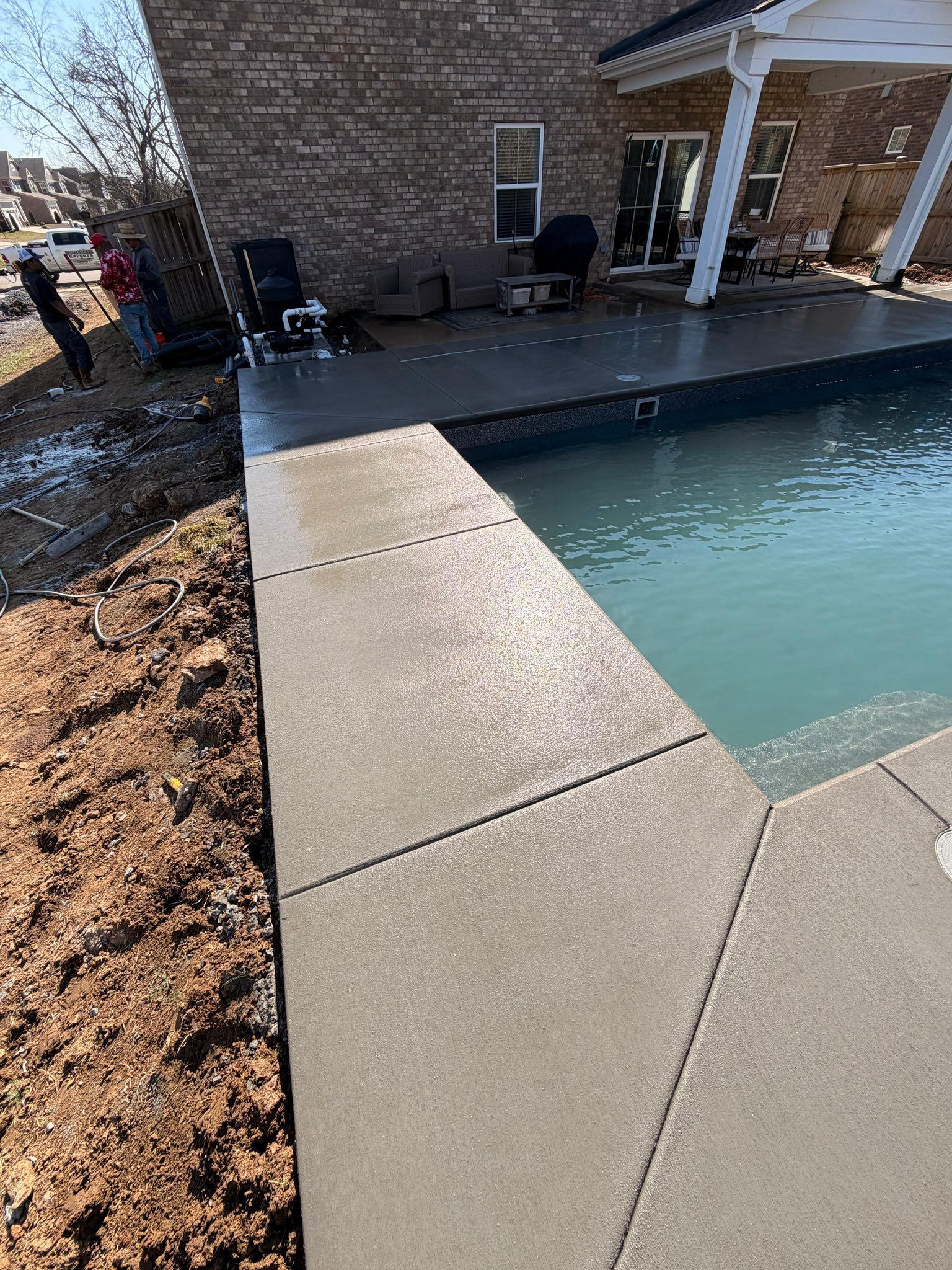 Concrete pool deck beside a turquoise in-ground pool next to a brick patio and house