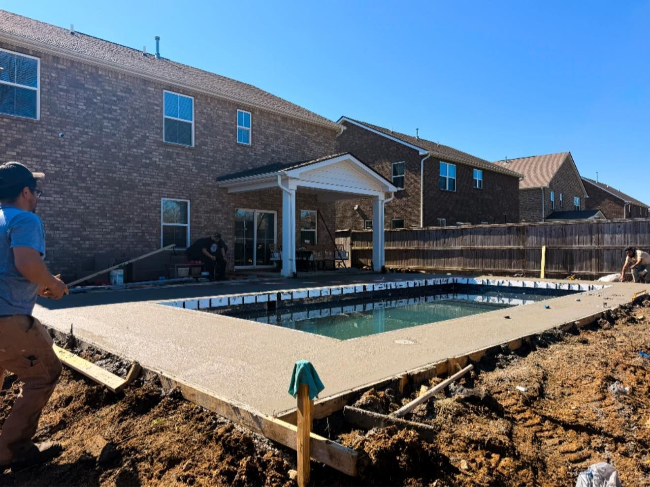Backyard under construction with a raised pool deck beside a brick house and workers nearby