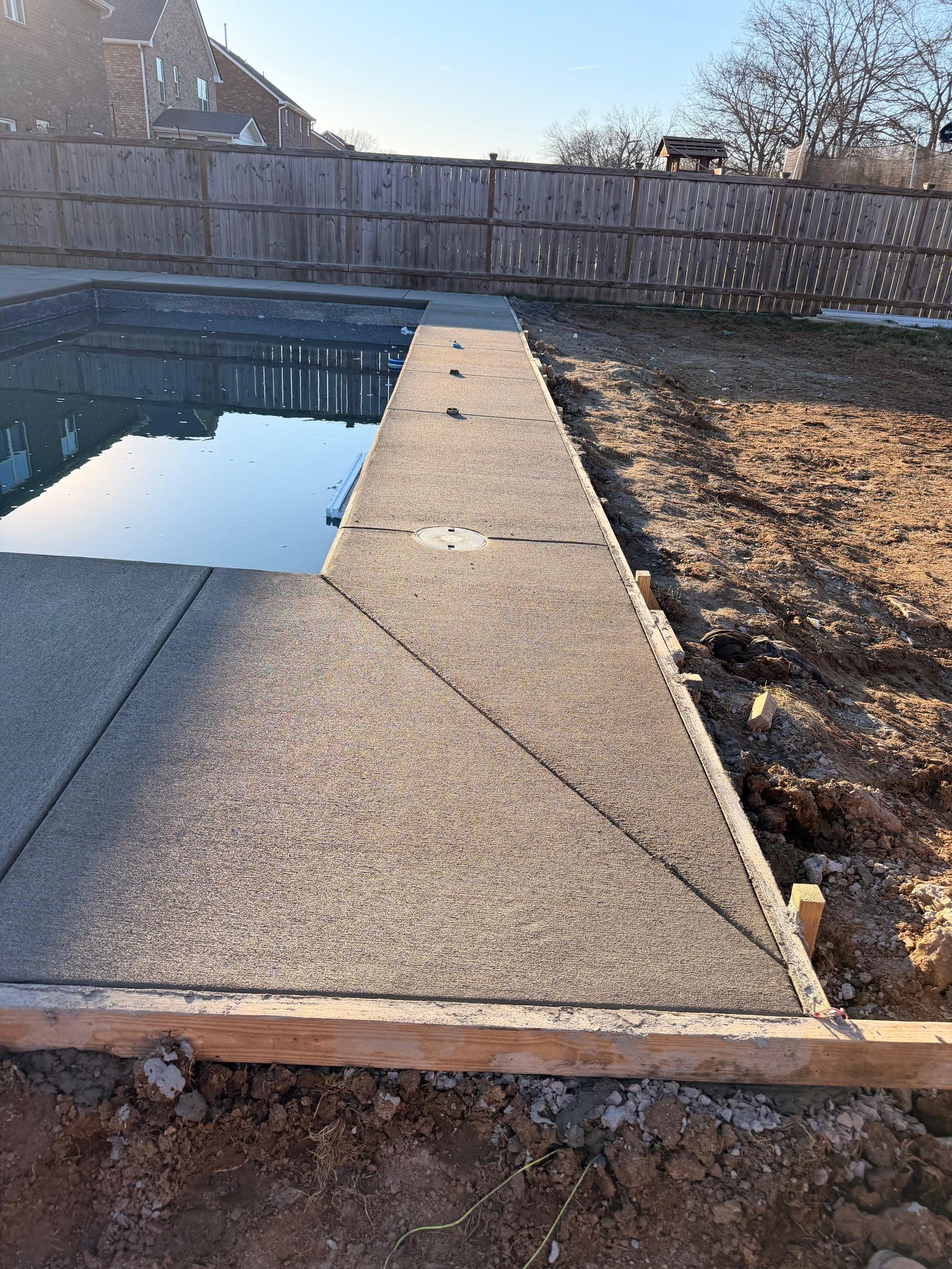 Backyard swimming pool with a concrete deck, wooden fence, and dry ground in bright sunlight