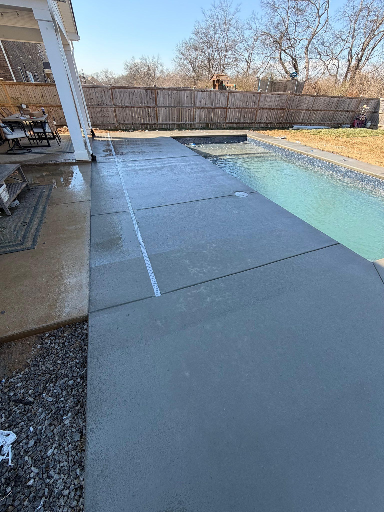 Backyard patio beside a swimming pool with gray concrete pavers and a wooden fence in the background