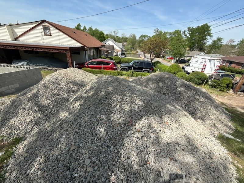 Large gravel pile in a residential driveway beside parked cars and houses on a sunny day