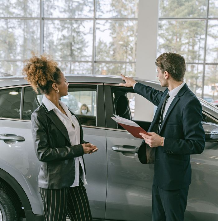 Two business people discussing a silver car in a bright showroom, one holding a clipboard.