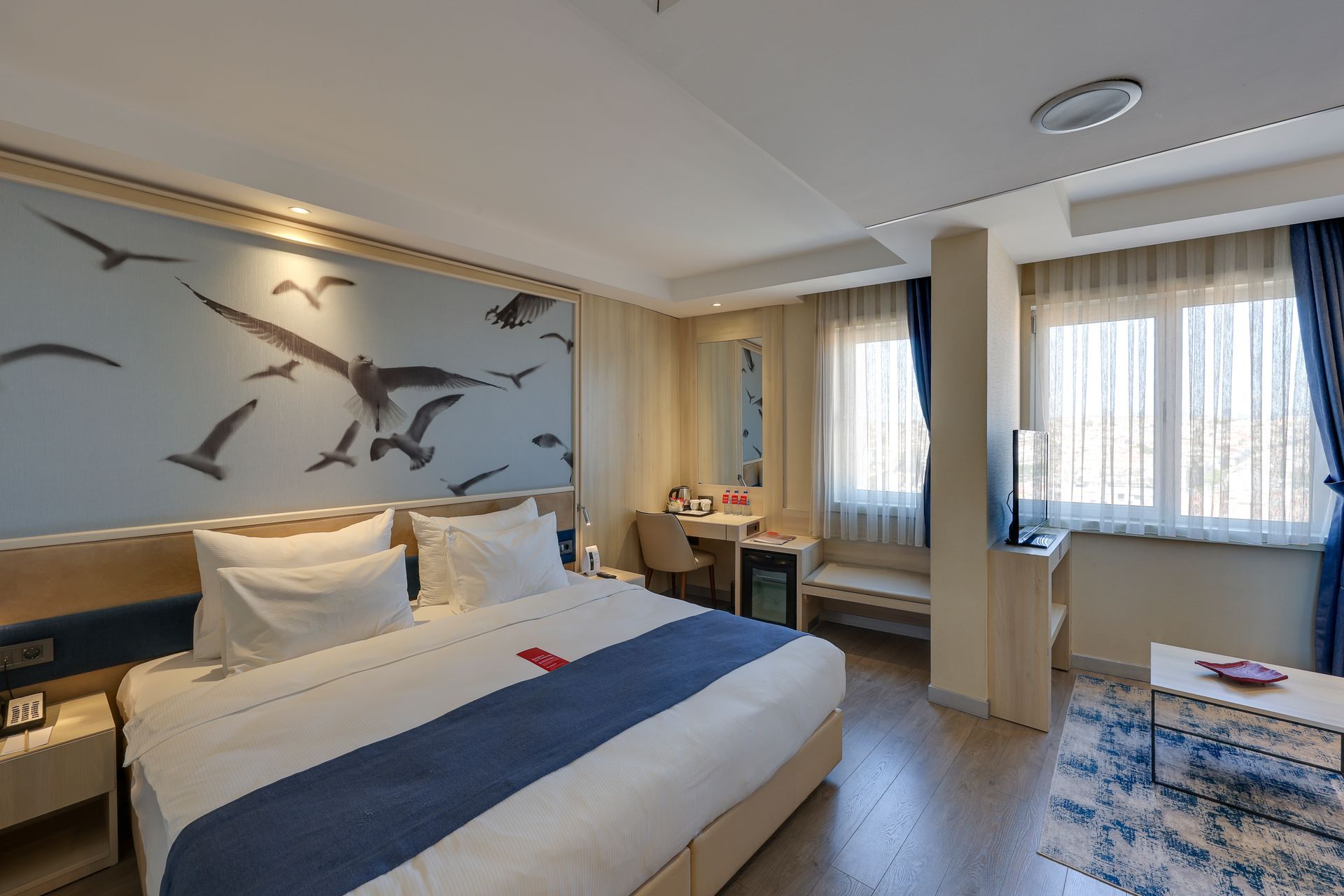 Hotel room with a large bed, seagull mural, desk, and two windows with blue curtains.